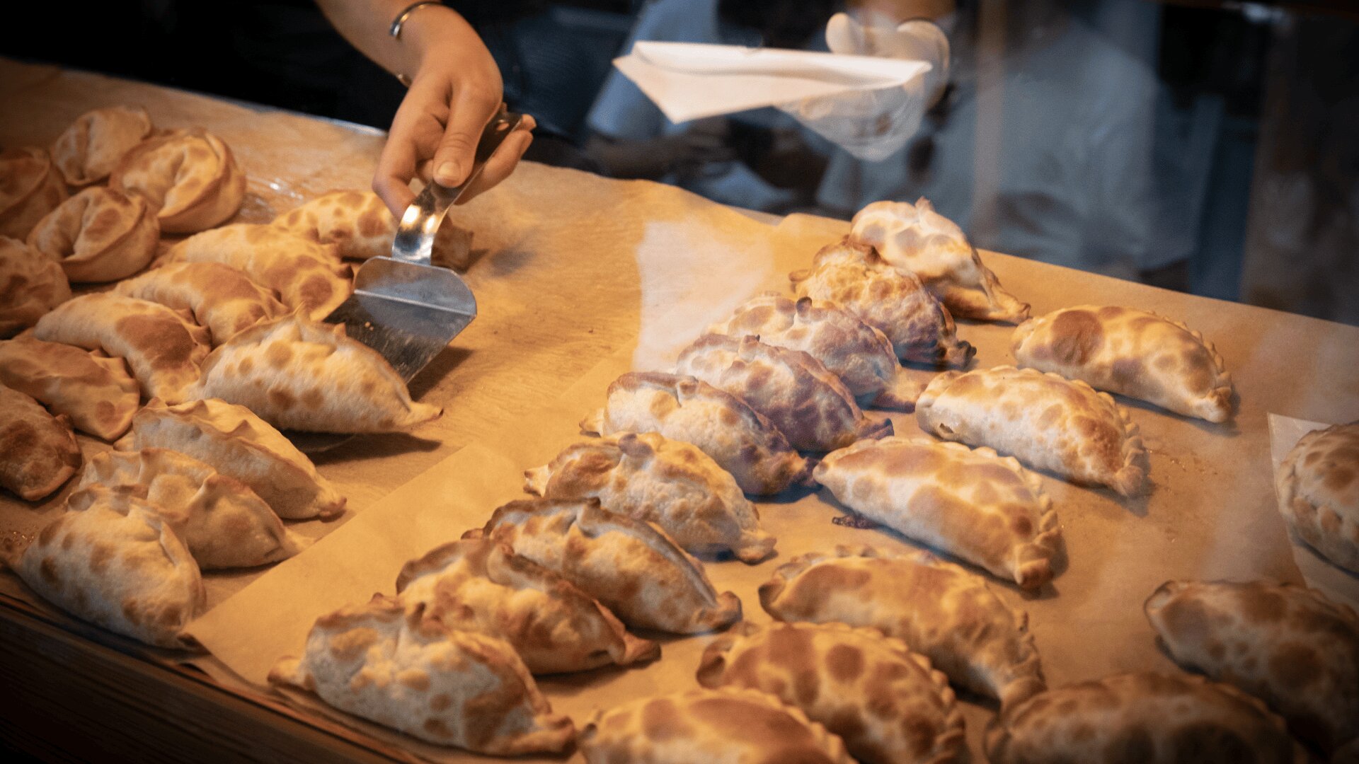 Eine Person nimmt mit einem Metallspatel eine goldbraune Empanada von einem Tablett, das mit vielen frisch gebackenen Empanadas auf Pergamentpapier gefüllt ist.