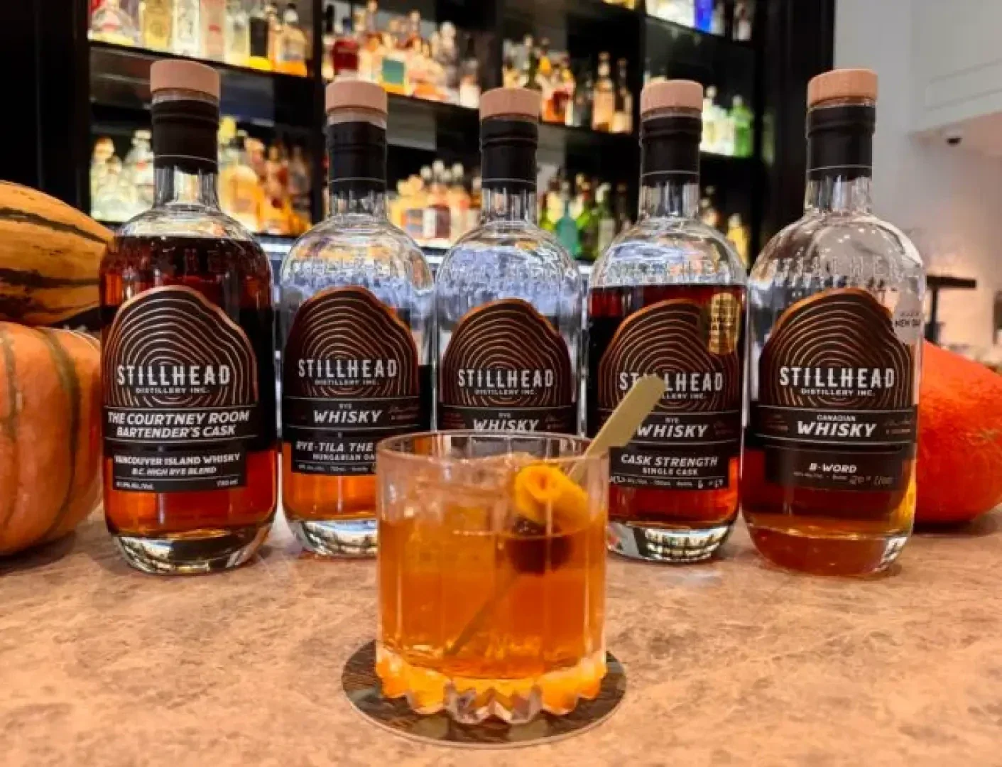 Fünf Flaschen Stillhead-Whisky sind auf einem Bartresen aufgereiht, vor dem ein mit Orangenschalen garnierter Cocktail steht. Im unscharfen Hintergrund sind Regale mit verschiedenen Flaschen zu sehen.
