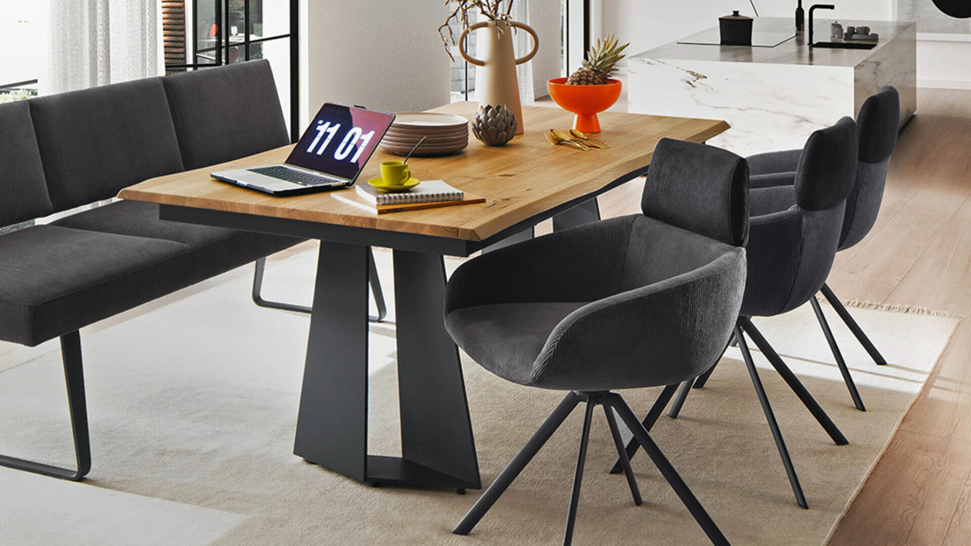 Ein moderner Essbereich mit einem Holztisch, schwarzen gepolsterten Stühlen, einer grauen Bank, einem Laptop, der die Uhrzeit anzeigt, einer gelben Tasse mit Untertasse, einem Notizbuch und dekorativen Gegenständen auf einem hellen Teppich in der Nähe großer Fenster.