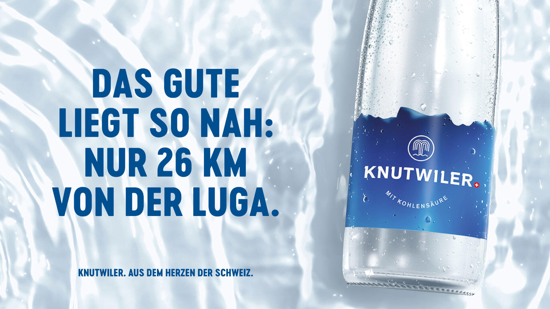 Eine klare Glasflasche mit Knutwiler Sprudel liegt auf einer nassen, eisigen Oberfläche. Ein grosser blauer Text in deutscher Sprache lautet: DAS GUTE LIEGT SO NAH: NUR 26 KM VON DER LUGA. Branding und Schweizer Herkunft sind hervorgehoben.