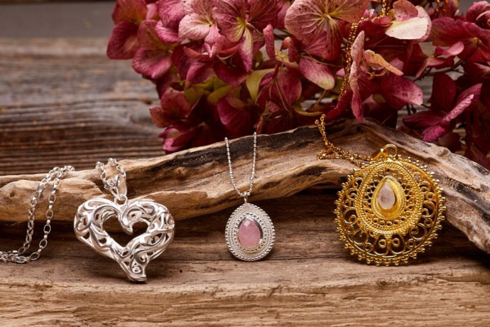 Drei verschnörkelte Anhänger - ein silbernes Herz, ein silbernes Oval mit einem rosafarbenen Stein und ein runder Goldanhänger - werden auf Holz mit getrockneten rosa Hortensienblüten im Hintergrund präsentiert.