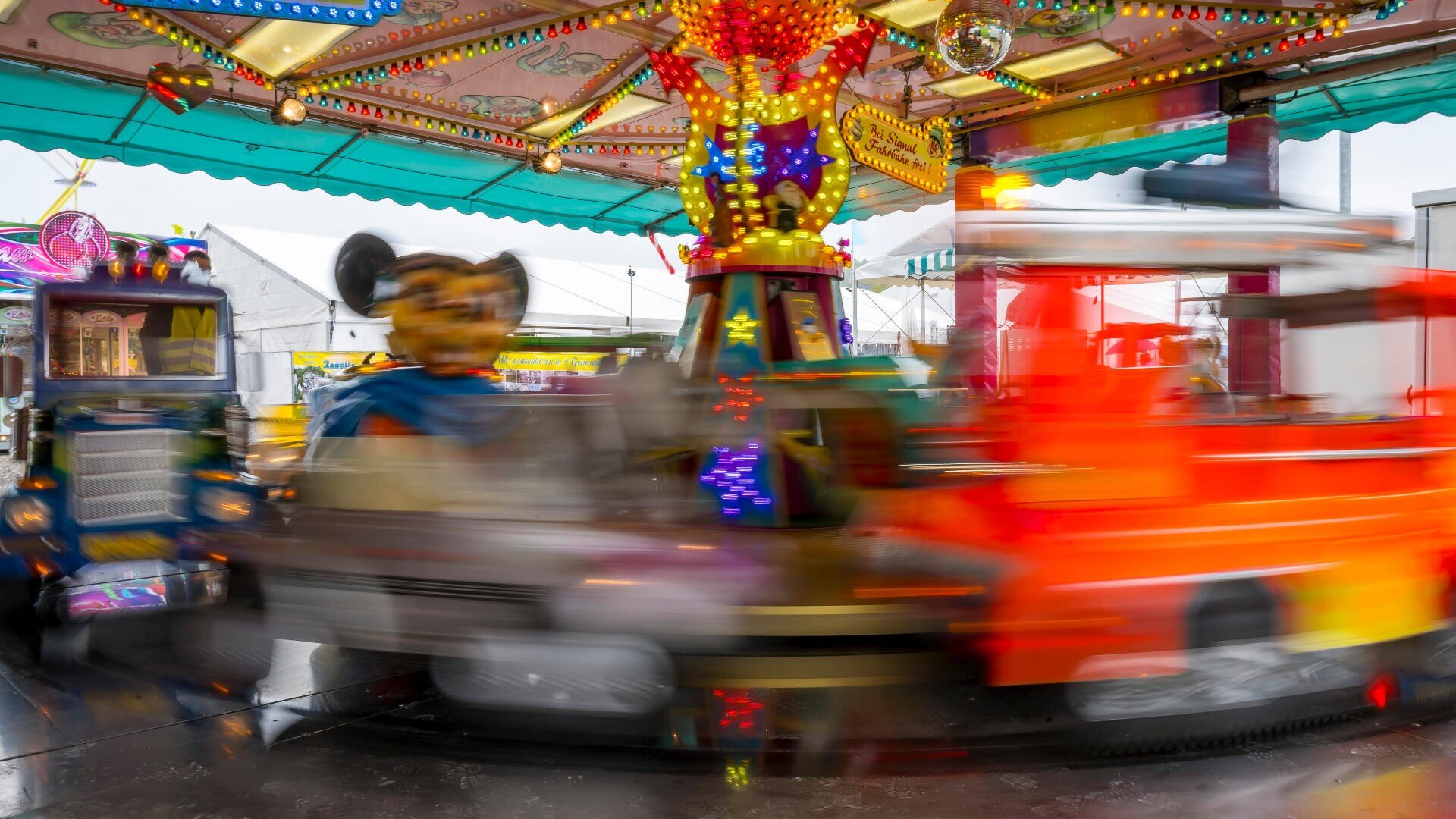 Ein hell beleuchtetes, farbenfrohes Karussell dreht sich auf einem Jahrmarkt, wobei verschwommene Spielzeugfahrzeuge und Zeichentrickfiguren, darunter eine, die einer Maus ähnelt, ein Gefühl von Bewegung und Aufregung erzeugen.
