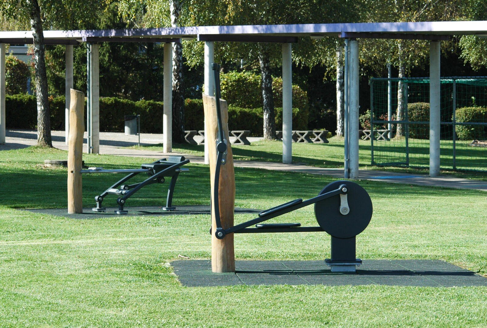 Die Fitnessgeräte im Freien stehen auf einer Wiese in einem Park, mit Bäumen, Bänken und einem überdachten Weg im Hintergrund. Die Fitnessgeräte sind schwarz mit Holzpfosten und stehen auf schwarzen Matten.