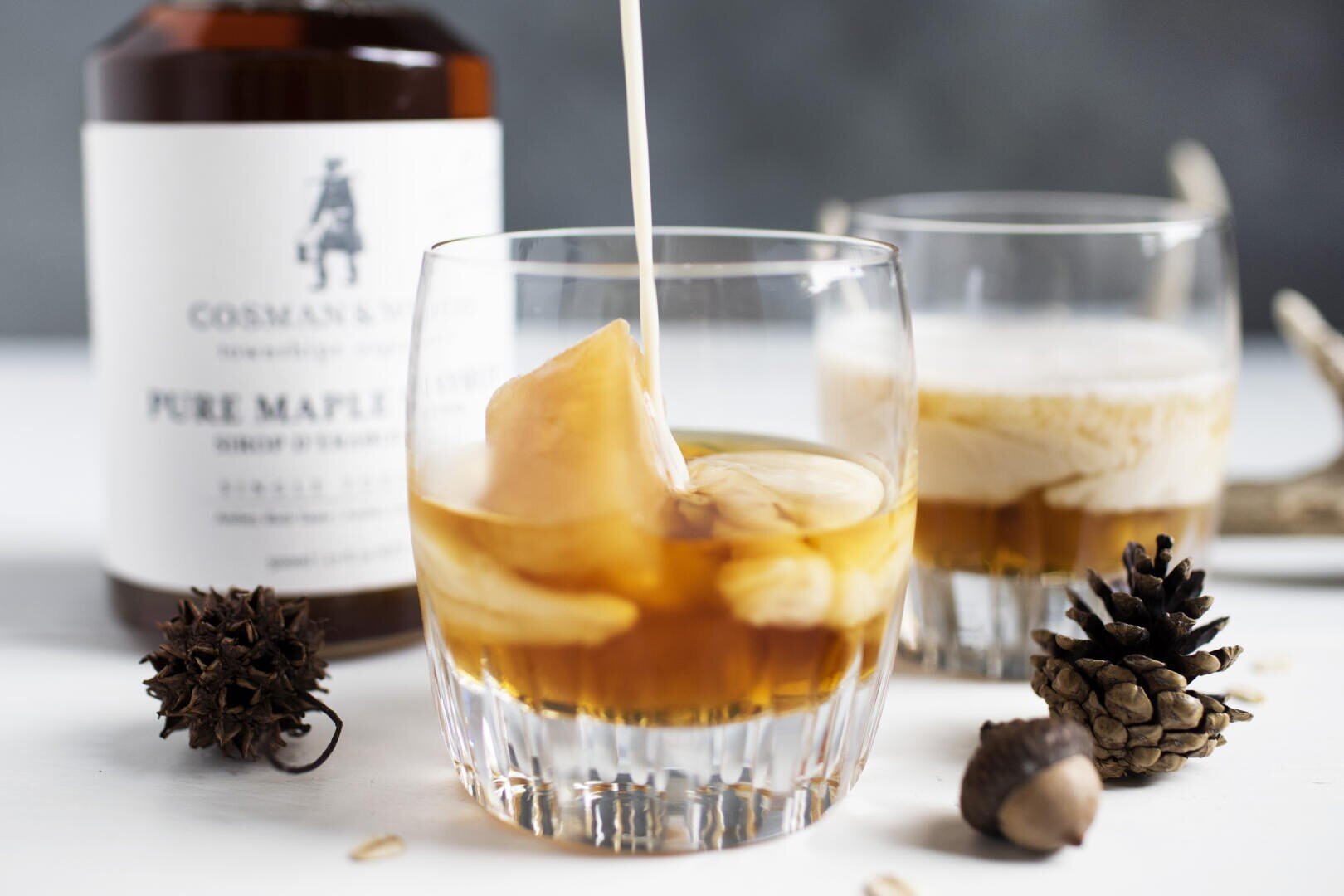 Ein Cocktailglas mit Eis wird mit Sahne aufgefüllt, daneben steht ein ähnliches Getränk. Auf dem Tisch stehen eine Flasche mit der Aufschrift Pure Maple und herbstliche Dekorationsartikel wie Tannenzapfen und eine Eichel.