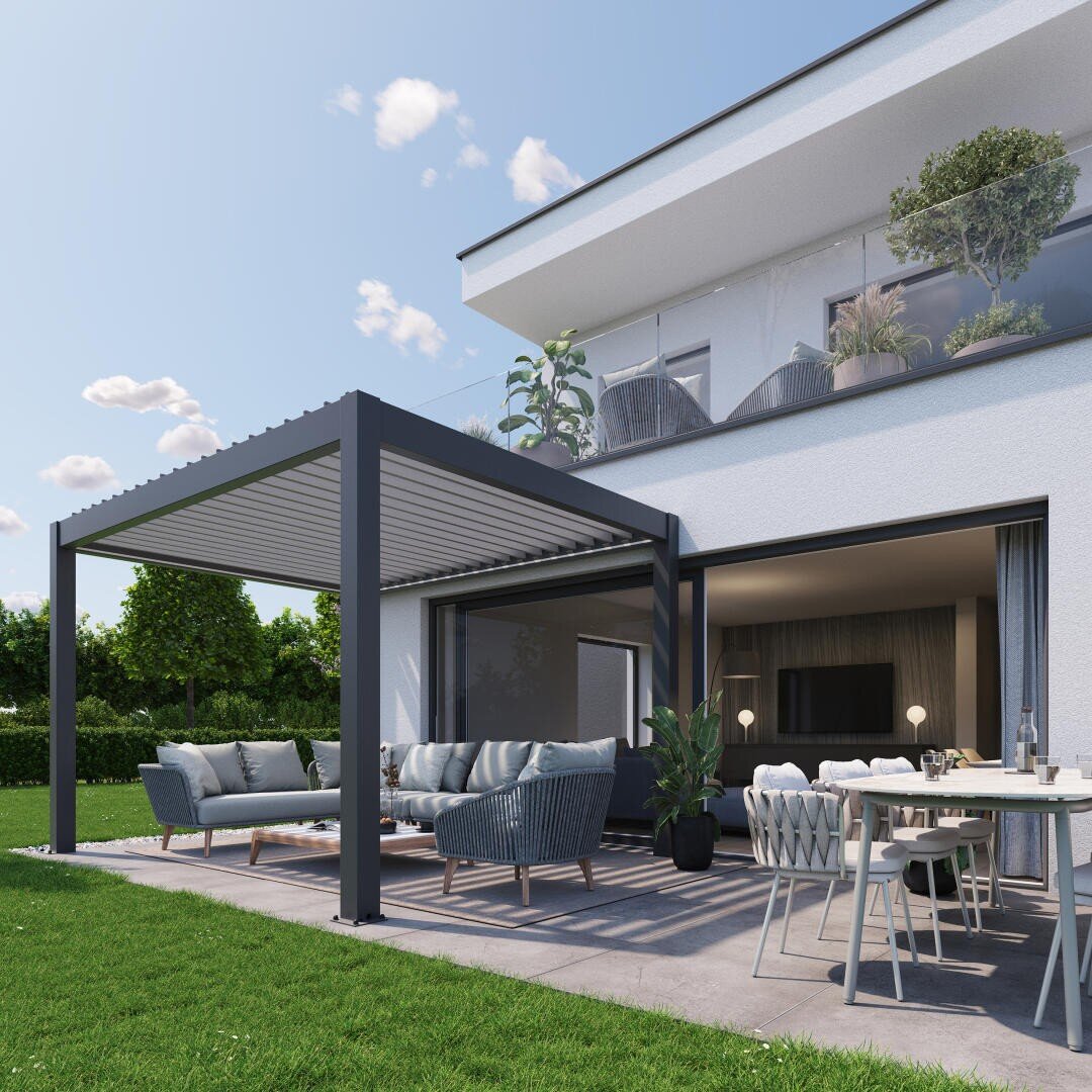Moderner Innenhof mit einer Pergola, Sofas und einem Esstisch auf einer Betonterrasse neben einem weißen Haus. Große Fenster öffnen sich zu einem modernen Wohnzimmer. Grüner Rasen und Bäume umgeben den Raum unter einem blauen Himmel mit Wolken.