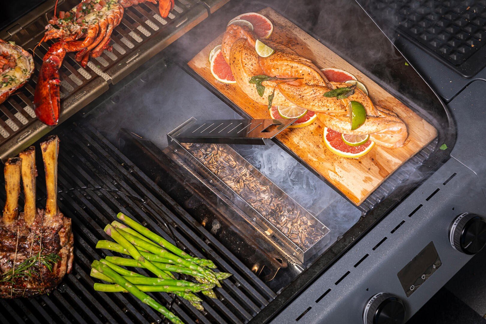 Lachsfilets mit Limettenscheiben werden auf einer Zedernholzplanke neben Spargel, Rippchen und Hummerhälften auf einem rauchigen Grill gegrillt. Holzspäne in einer Räucherbox sorgen für zusätzlichen Geschmack.