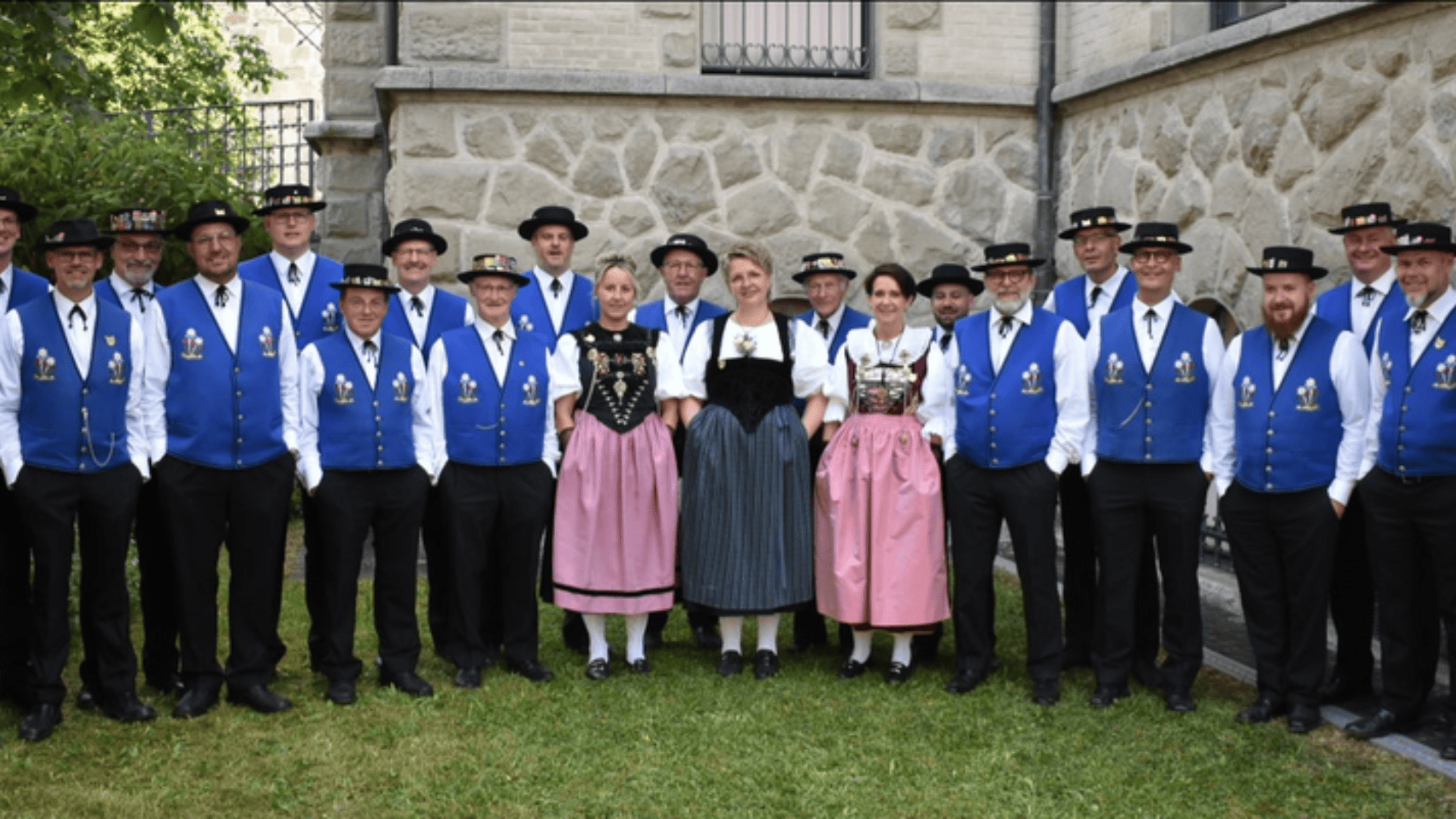 Eine Gruppe von Männern und Frauen posiert im Freien in traditioneller deutscher Kleidung, wobei die Männer blaue Westen und Hüte und die Frauen Dirndl tragen und vor einem Steingebäude stehen.