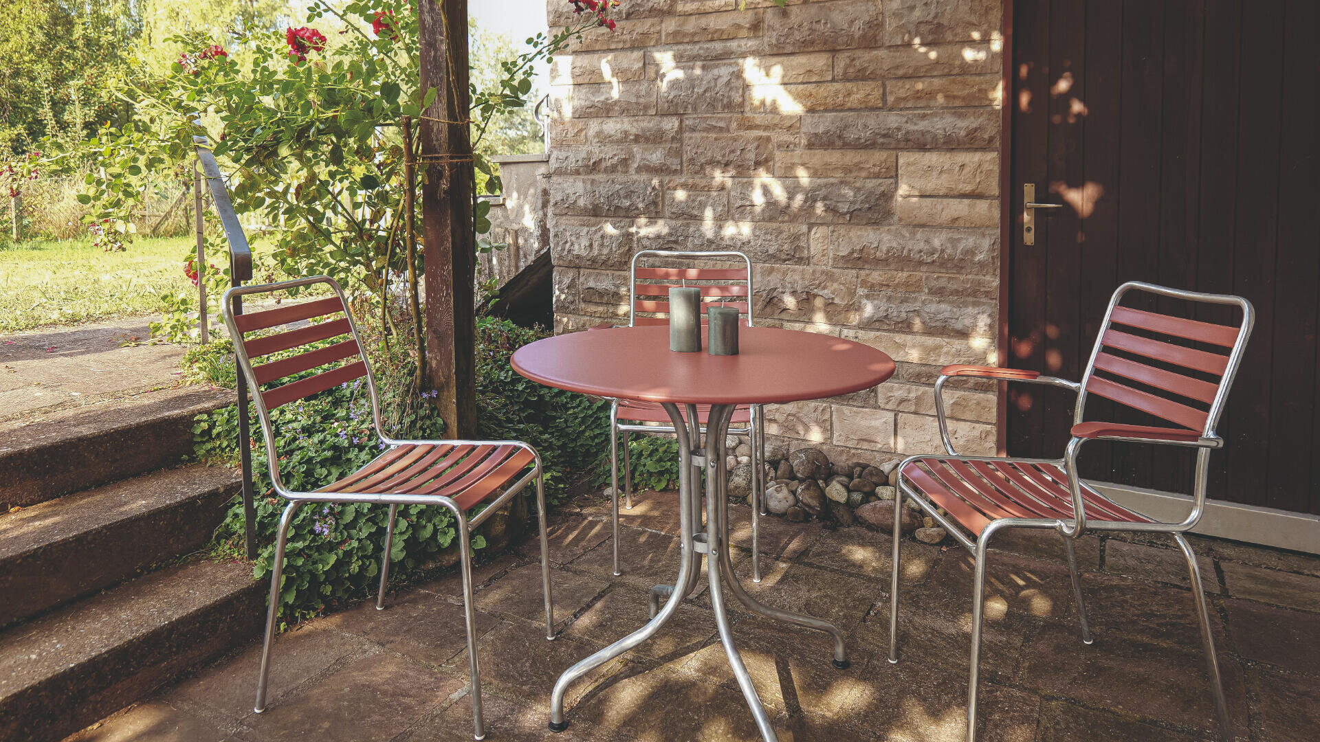 Ein runder, roter Tisch mit drei passenden roten und silbernen Stühlen steht auf einer Terrasse in der Nähe einer Backsteinmauer und einer Tür. Das Sonnenlicht dringt durch das Laub und wirft schräge Schatten auf die Szene.