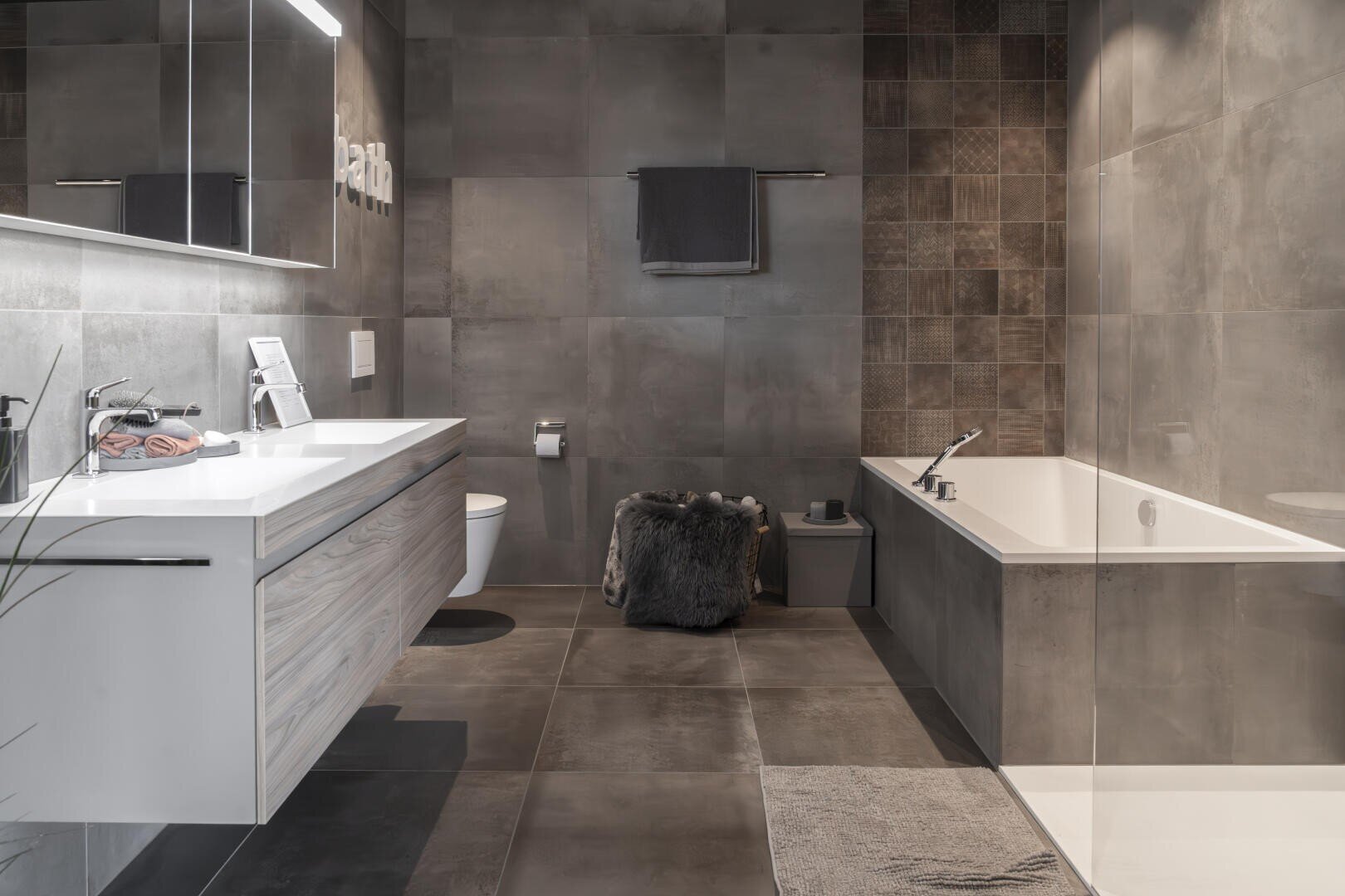 Modernes Badezimmer mit grauen Fliesen, schwebendem Waschtisch mit Doppelwaschbecken, wandmontierter Toilette, Badewanne, Glasdusche und einem Hocker mit Fell. Minimalistisches Dekor mit sanfter Beleuchtung und neutralen Tönen.