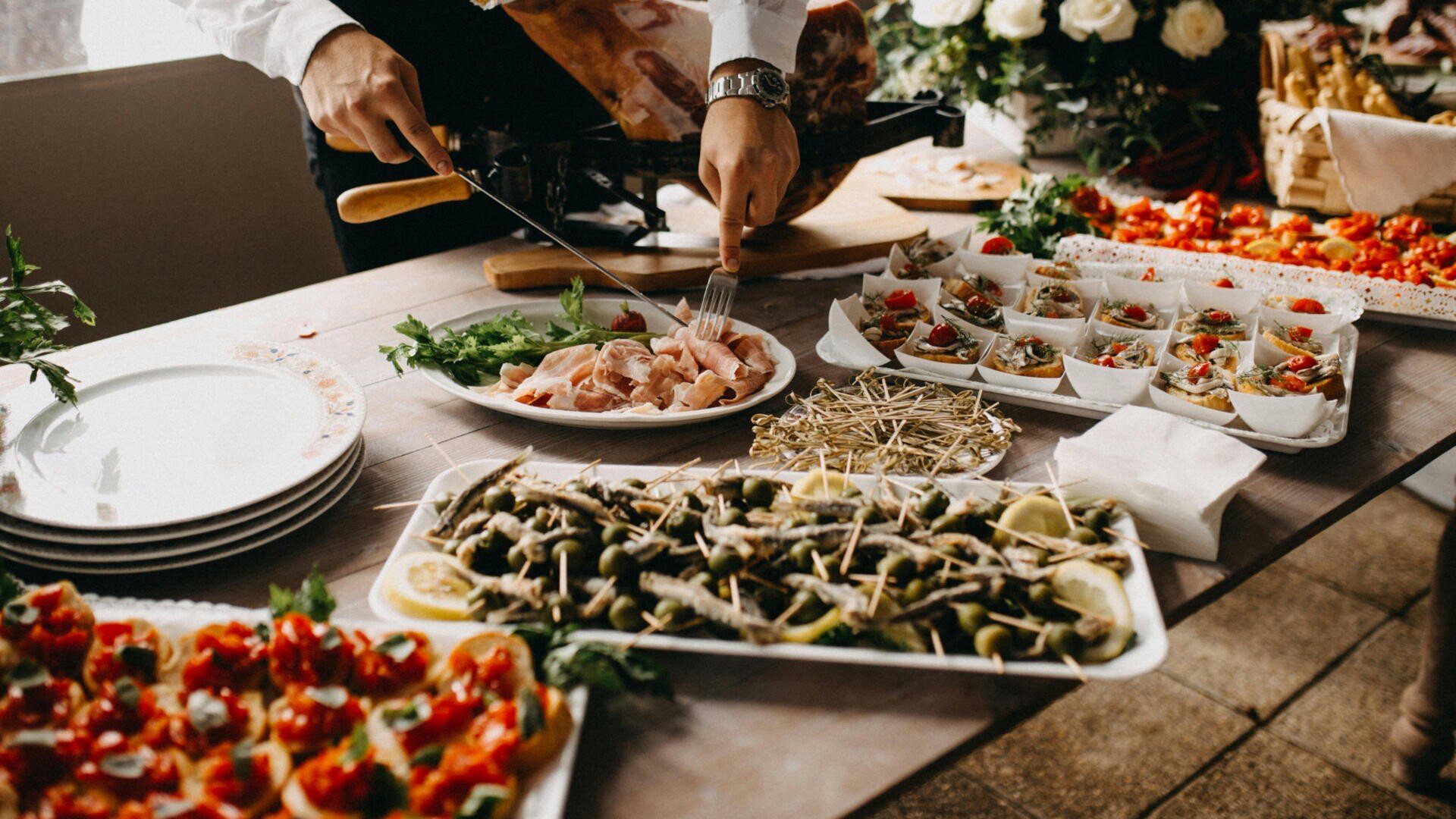 Eine Person serviert das Essen von einem Buffettisch mit verschiedenen Vorspeisen, darunter Fleisch, Gemüse und Salate, die auf Platten und Schalen angerichtet sind, während sich in der Nähe Teller stapeln und im Hintergrund Blumen zu sehen sind.