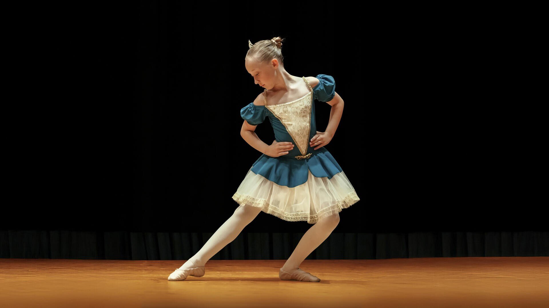 Eine junge Balletttänzerin in einem blau-weißen Kostüm tritt auf der Bühne auf. Sie blickt nach unten, stützt die Hände auf die Hüften und zeigt mit einem Fuß zur Seite, vor einem schwarzen Hintergrund.