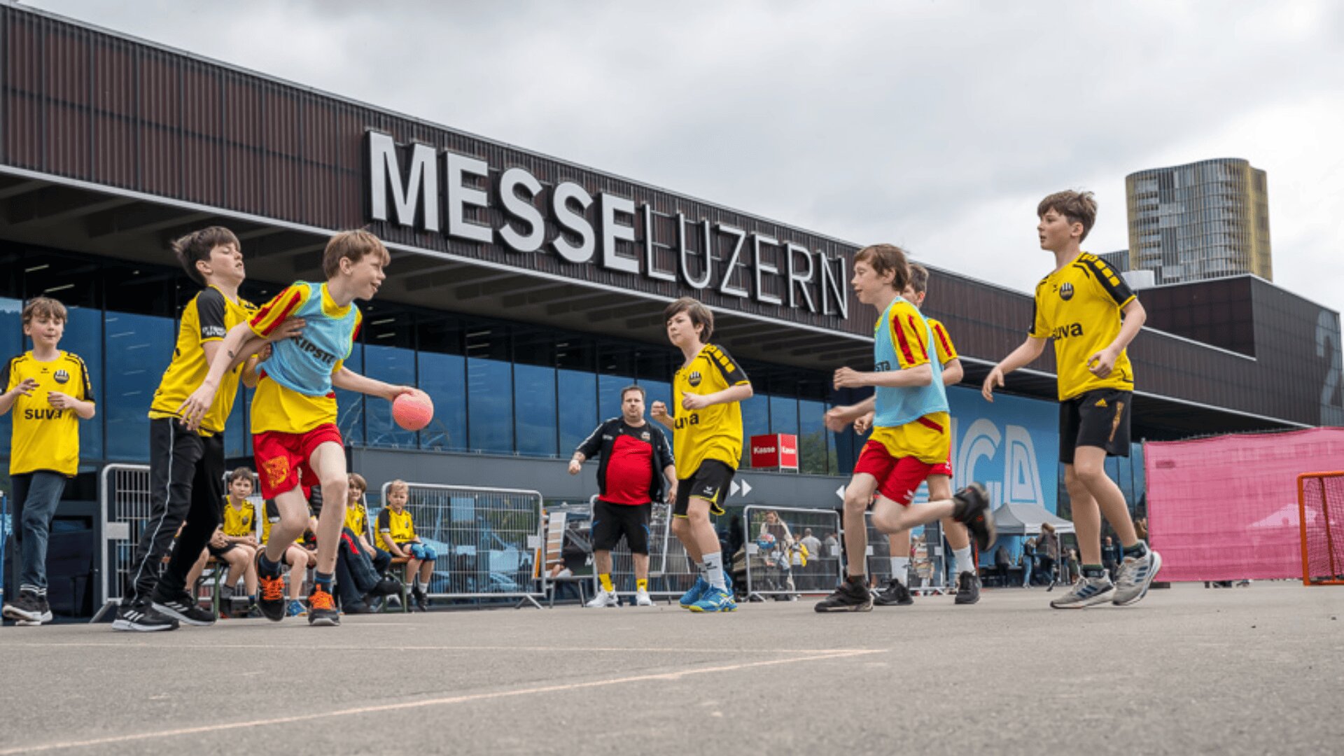 Eine Gruppe von Kindern in gelben Trikots spielt vor dem Kongresszentrum der Messe Luzern ein Ballspiel, während die Erwachsenen in der Nähe bei bewölktem Himmel zusehen.