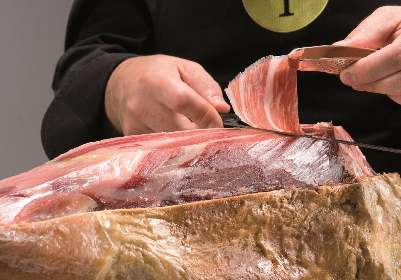 Eine Person schneidet mit einem scharfen Messer ein dünnes Stück Rohschinken aus einer großen Schinkenkeule und hält das Fleisch mit einer Hand fest.