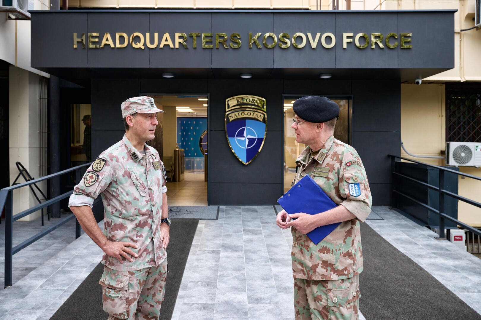 Zwei uniformierte Soldaten stehen vor dem Eingang zum Gebäude des Hauptquartiers der Kosovo-Truppen und unterhalten sich, während hinter ihnen NATO- und KFOR-Schilder zu sehen sind. Einer hält eine blaue Mappe in der Hand, beide tragen Tarnuniformen.
