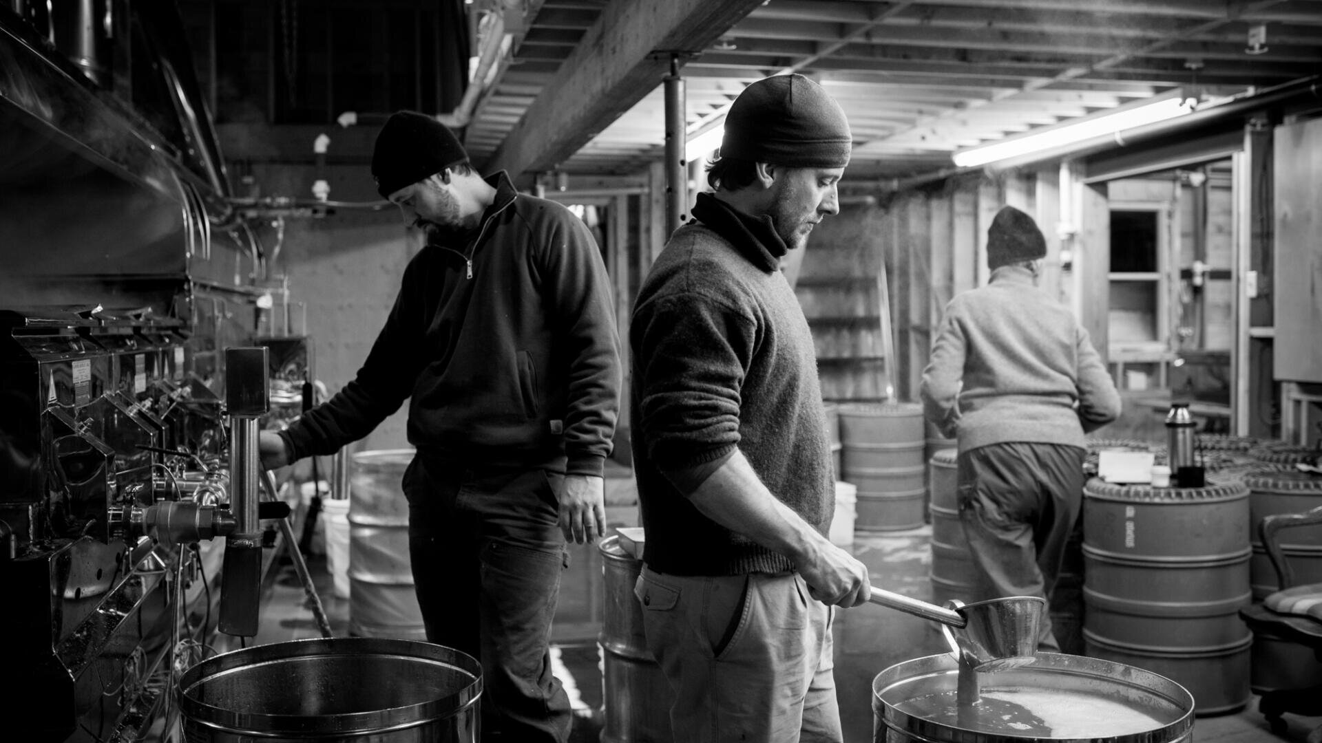 Drei Personen arbeiten in einer rustikalen, industriellen Umgebung. Zwei Männer im Vordergrund tragen Strickmützen und Pullover und kümmern sich um große Metallfässer. Im Hintergrund arbeitet eine weitere Person in der Nähe gestapelter Fässer. Die Szene ist in schwarz-weiß gehalten.