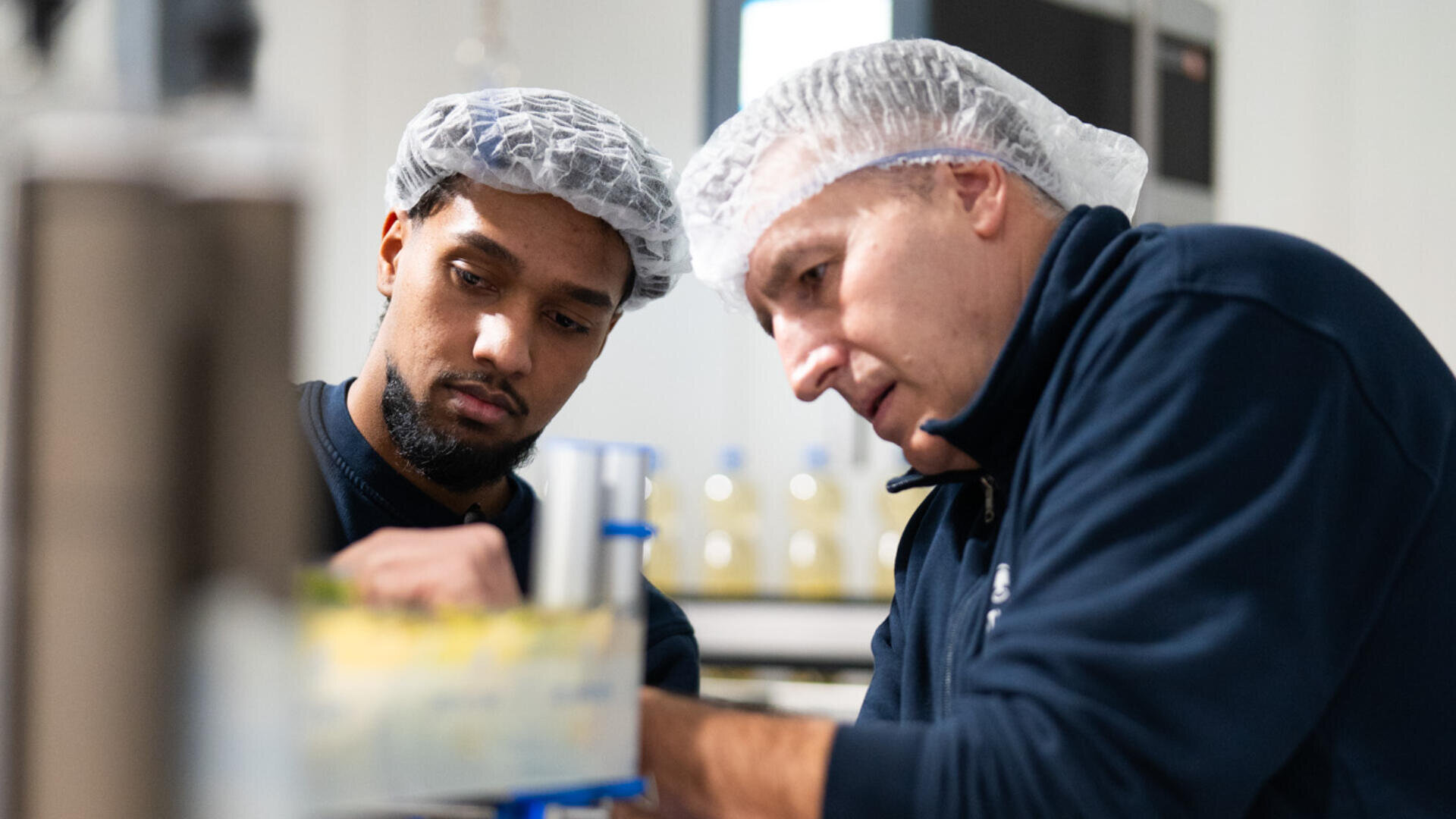Zwei Männer mit Haarnetzen und dunklen Uniformen beobachten und arbeiten an einer Maschine oder einem Gerät in einer scheinbaren Fabrik- oder Laborumgebung. Im unscharfen Hintergrund sind Flaschen zu sehen.