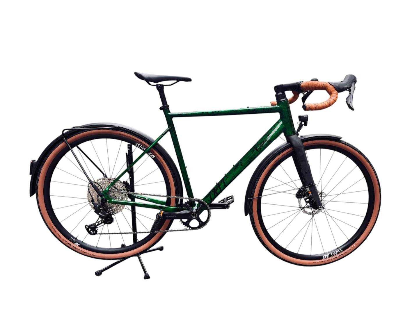 Ein grünes Schotterfahrrad mit braunem Lenkerband und passenden braunen Reifen ist auf einem weißen Hintergrund abgebildet. Das Fahrrad hat volle Schutzbleche, Scheibenbremsen und einen Ständer.