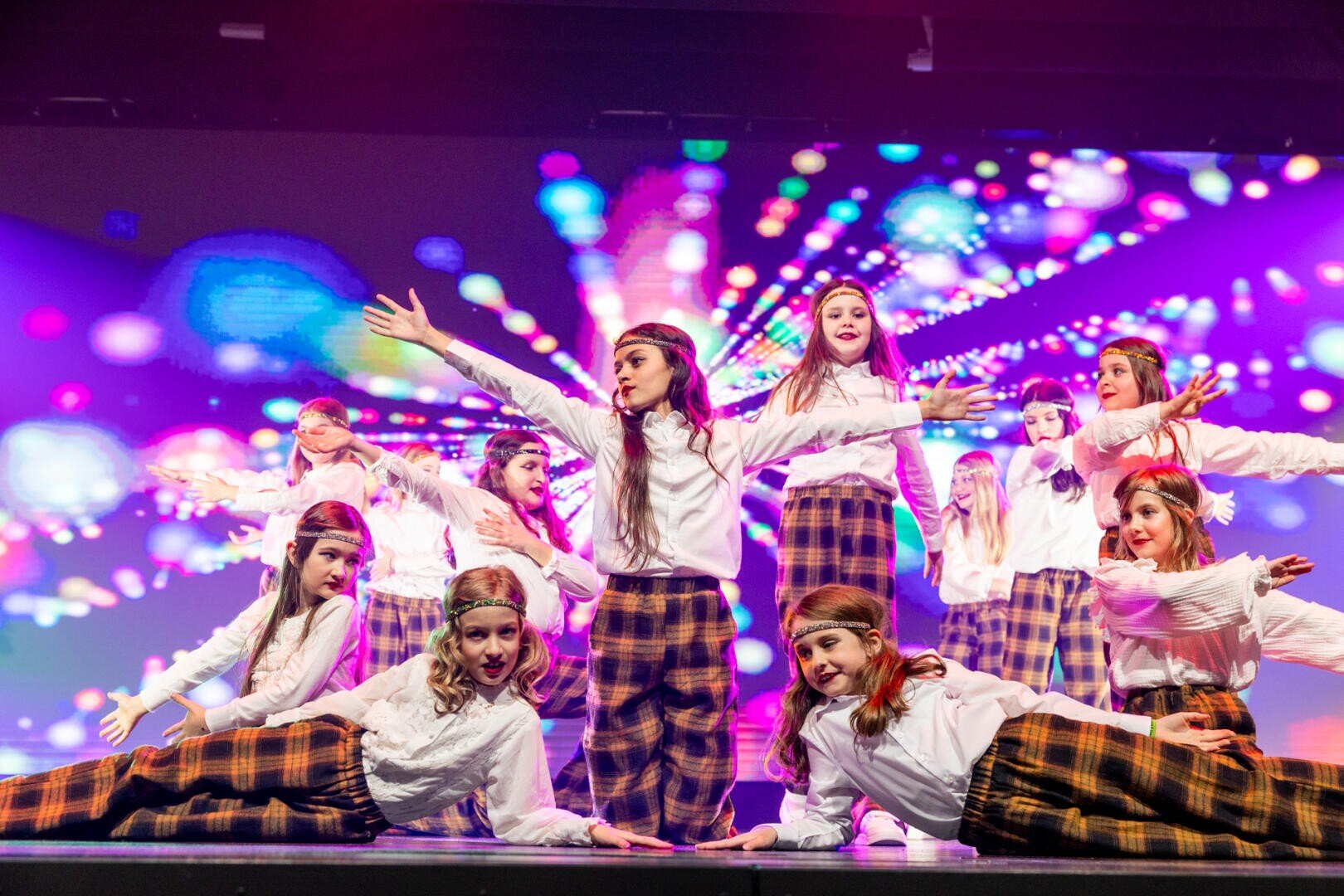 Eine Gruppe von Mädchen in karierten Hosen, weißen Hemden und Stirnbändern führt auf der Bühne einen choreografierten Tanz vor bunten, leuchtenden Lichtern und Mustern im Hintergrund auf. Einige haben die Arme ausgestreckt, während andere auf dem Boden posieren.