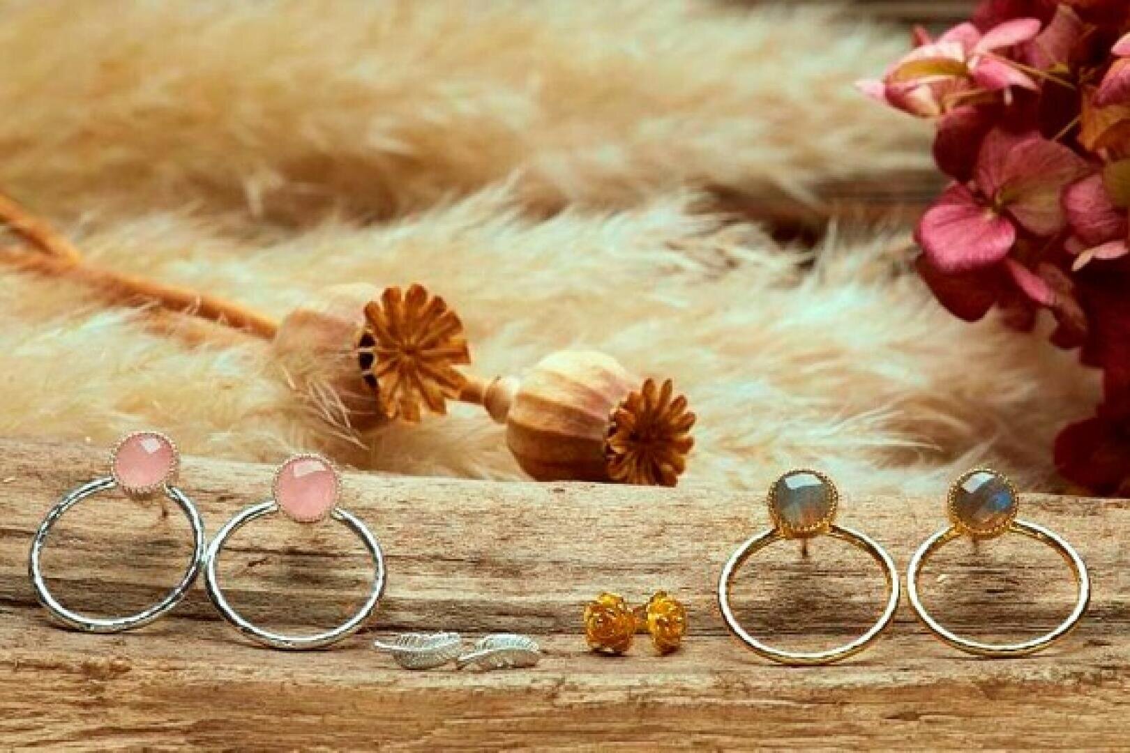 Eine Auslage mit verschiedenen Ohrringen auf einer rustikalen Holzfläche, darunter silberne und goldene Ohrringe mit Edelsteinen, kleine goldene Ohrstecker, silberne blattförmige Ohrstecker und rosa Blumen und getrocknete Pflanzen im Hintergrund.