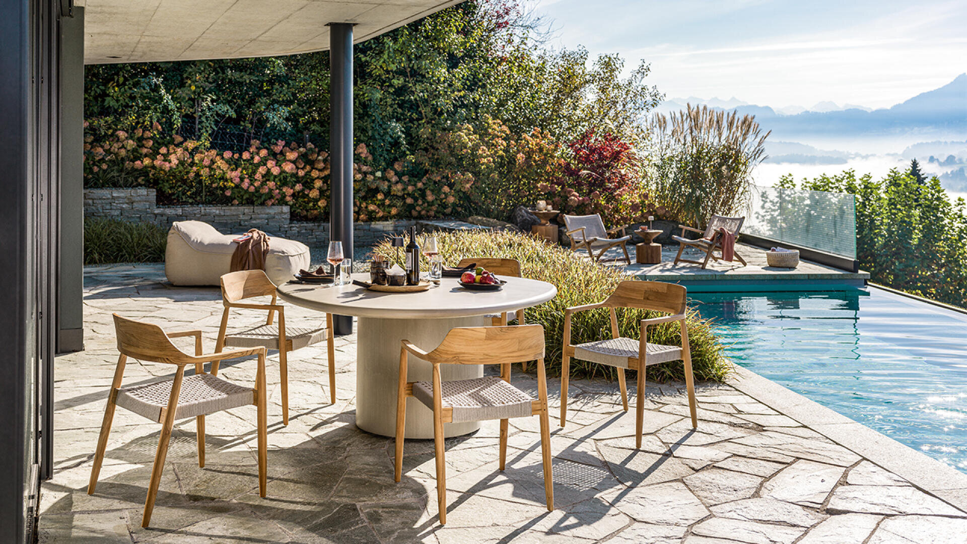 Auf der modernen Außenterrasse steht ein runder Tisch mit Getränken und Snacks, umgeben von vier Holzstühlen, neben einem Pool mit Liegestühlen, üppigen Pflanzen und einem malerischen Bergblick im Hintergrund.