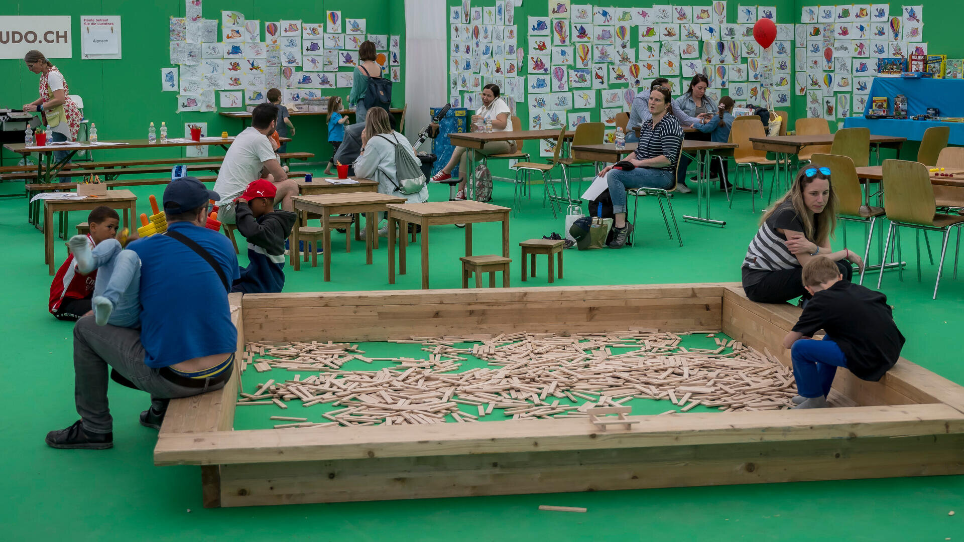 Kinder und Erwachsene befinden sich in einem Spielzimmer mit grünen Böden und Wänden, sitzen an Tischen und einem großen hölzernen Sandkasten, der mit Holzstöcken gefüllt ist. An den Wänden sind bunte Zeichnungen zu sehen.
