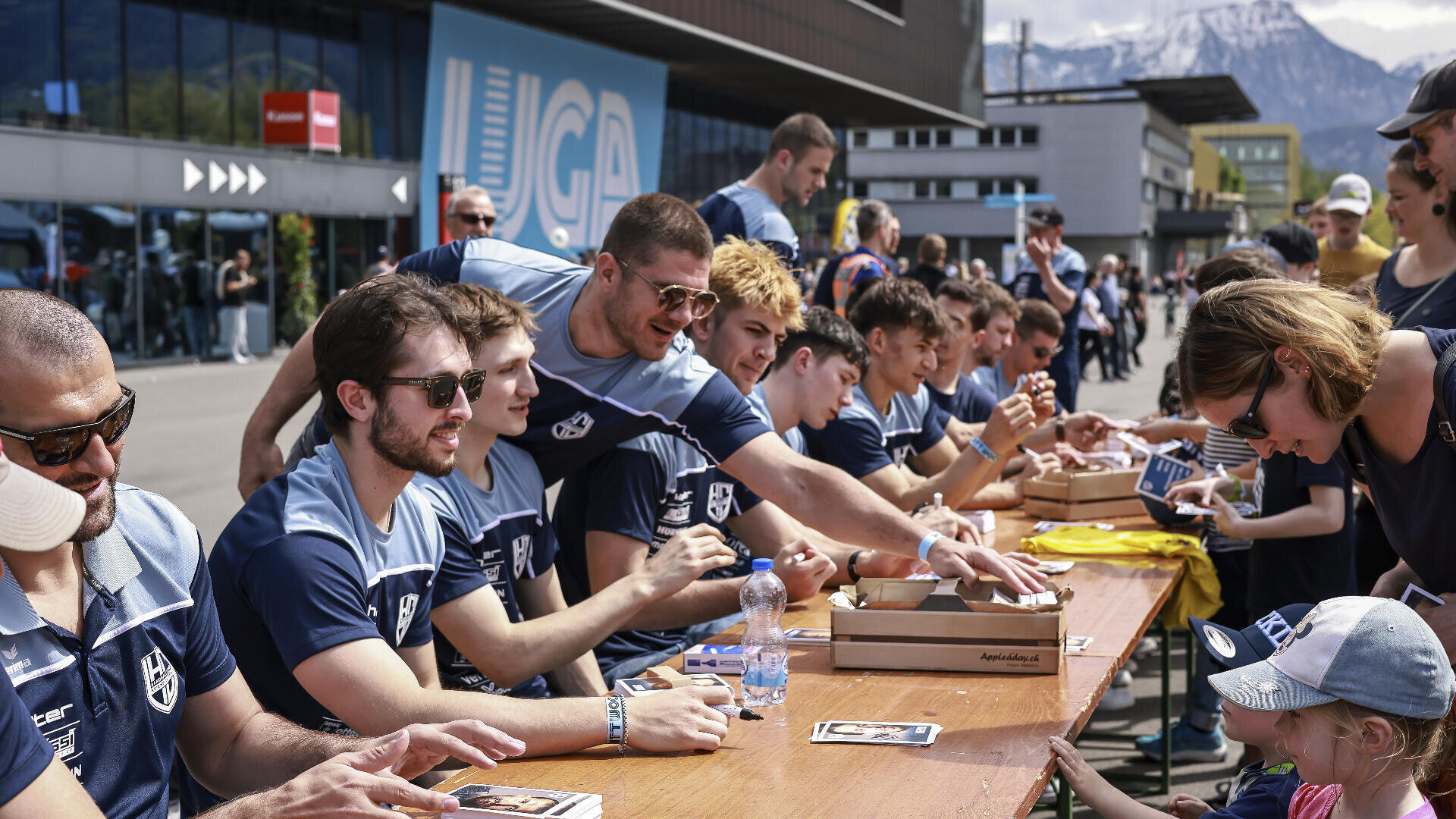 Eine Gruppe von Sportlern in passenden blauen Trikots sitzt an einem langen Tisch im Freien und gibt den Fans Autogramme, während im Hintergrund Berge und moderne Gebäude zu sehen sind. Die Menschen auf beiden Seiten unterhalten sich und tauschen Gegenstände aus.