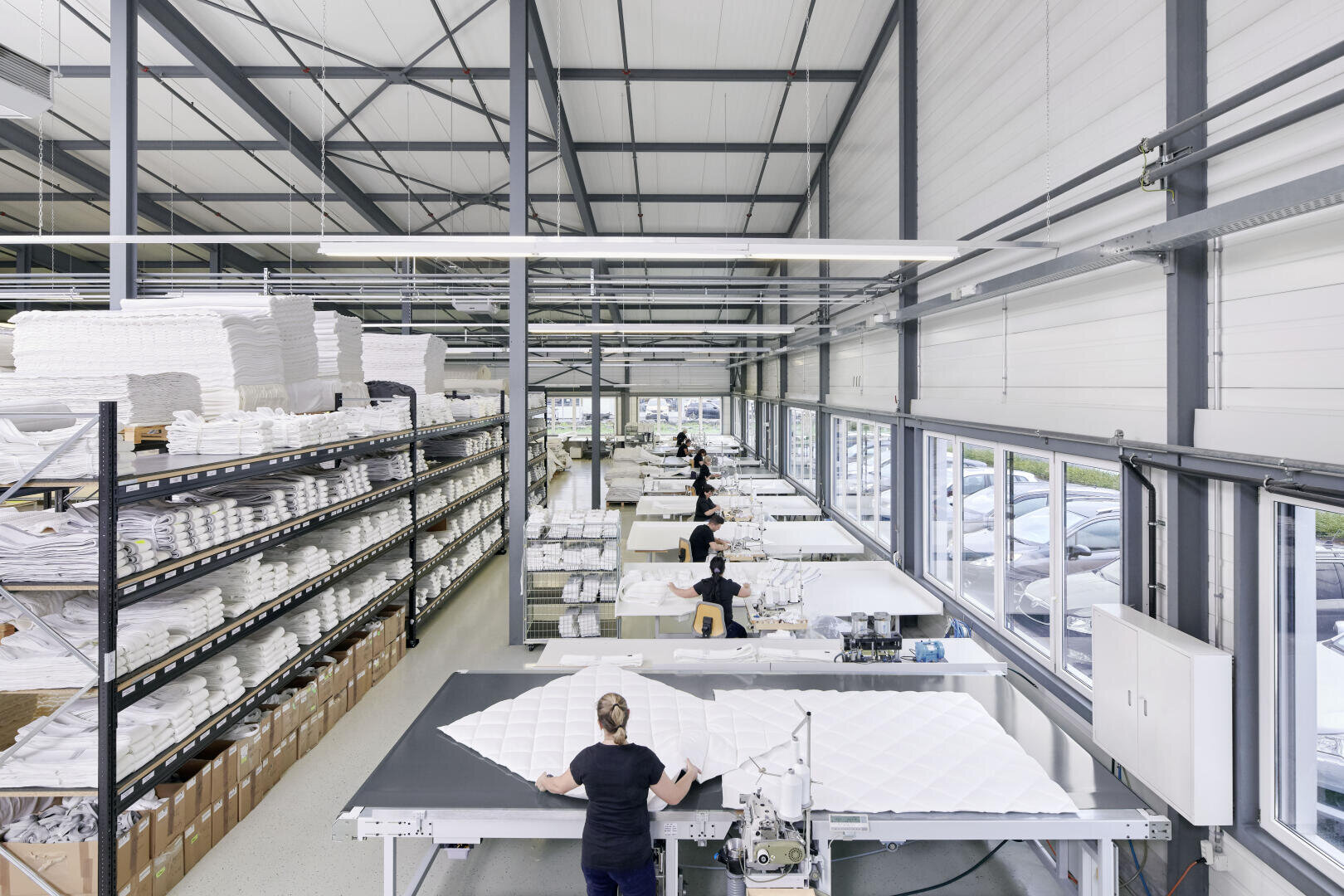 Arbeiter in einer hellen, gut organisierten Textilfabrik nähen und montieren große weiße Stoffstücke an langen Tischen. In den Regalen auf der linken Seite werden gestapelte weiße Stoffe aufbewahrt, und die großen Fenster auf der rechten Seite geben den Blick auf geparkte Autos frei.