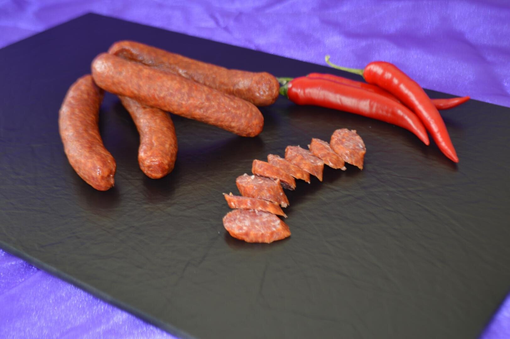 Mehrere ganze und in Scheiben geschnittene geräucherte Würste sind auf einer schwarzen Schieferplatte neben zwei ganzen roten Chilischoten angeordnet, mit einem lila Tuch im Hintergrund.