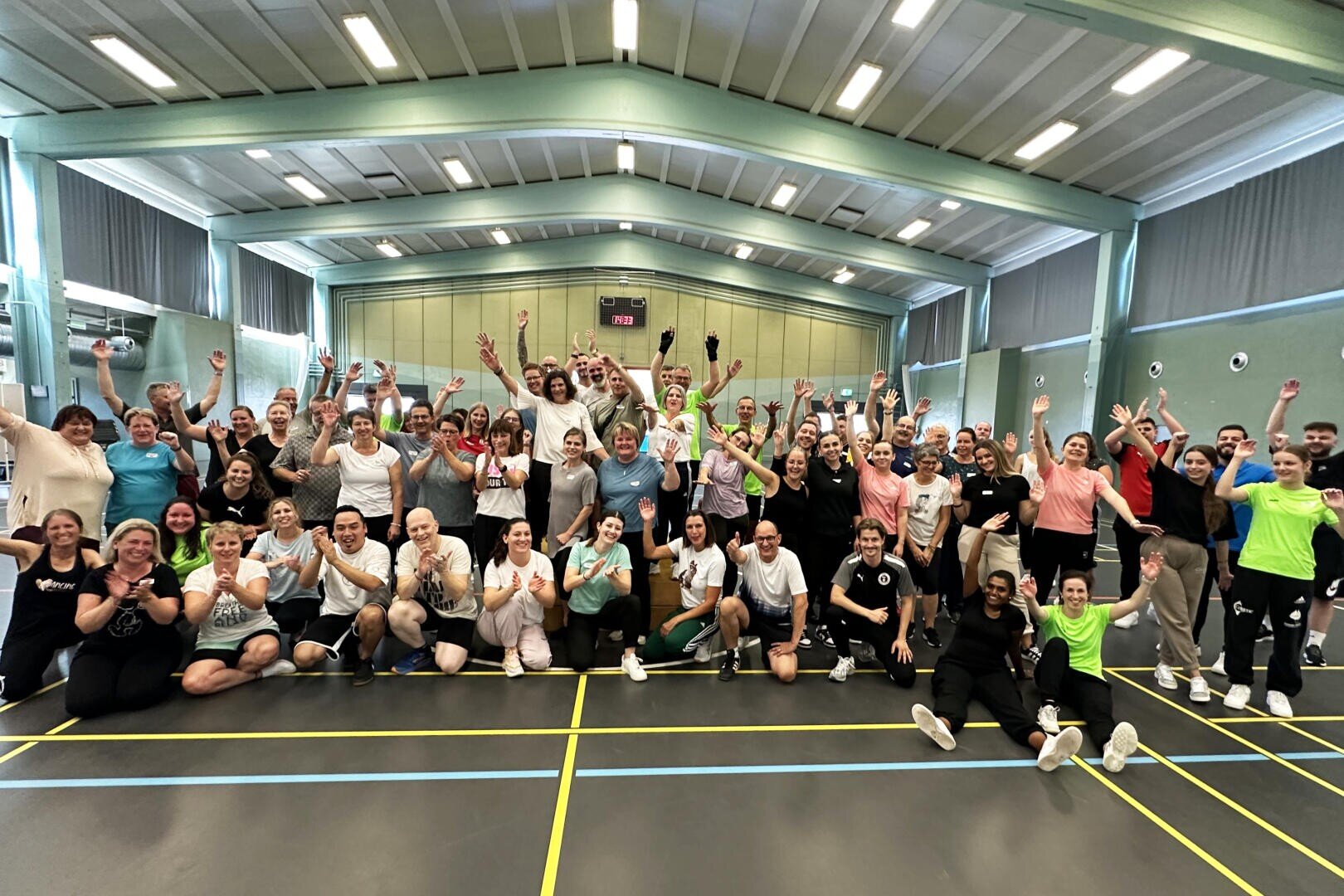 Eine große Gruppe von Menschen in Sportkleidung posiert gemeinsam auf einem Hallenplatz, lächelt und hebt feierlich die Arme. Zu der Gruppe gehören sowohl Männer als auch Frauen, und die Atmosphäre ist lebhaft und fröhlich.