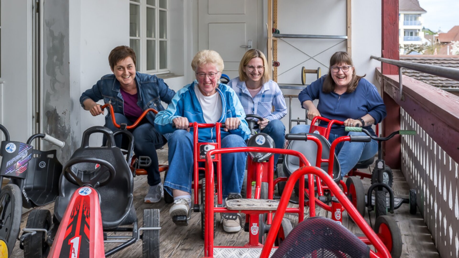 Vier lächelnde Erwachsene sitzen auf roten Dreirädern auf einem hölzernen Balkon und scheinen miteinander zu rasen und Spaß zu haben. Sie sind im Freien, und die Atmosphäre ist spielerisch und fröhlich.