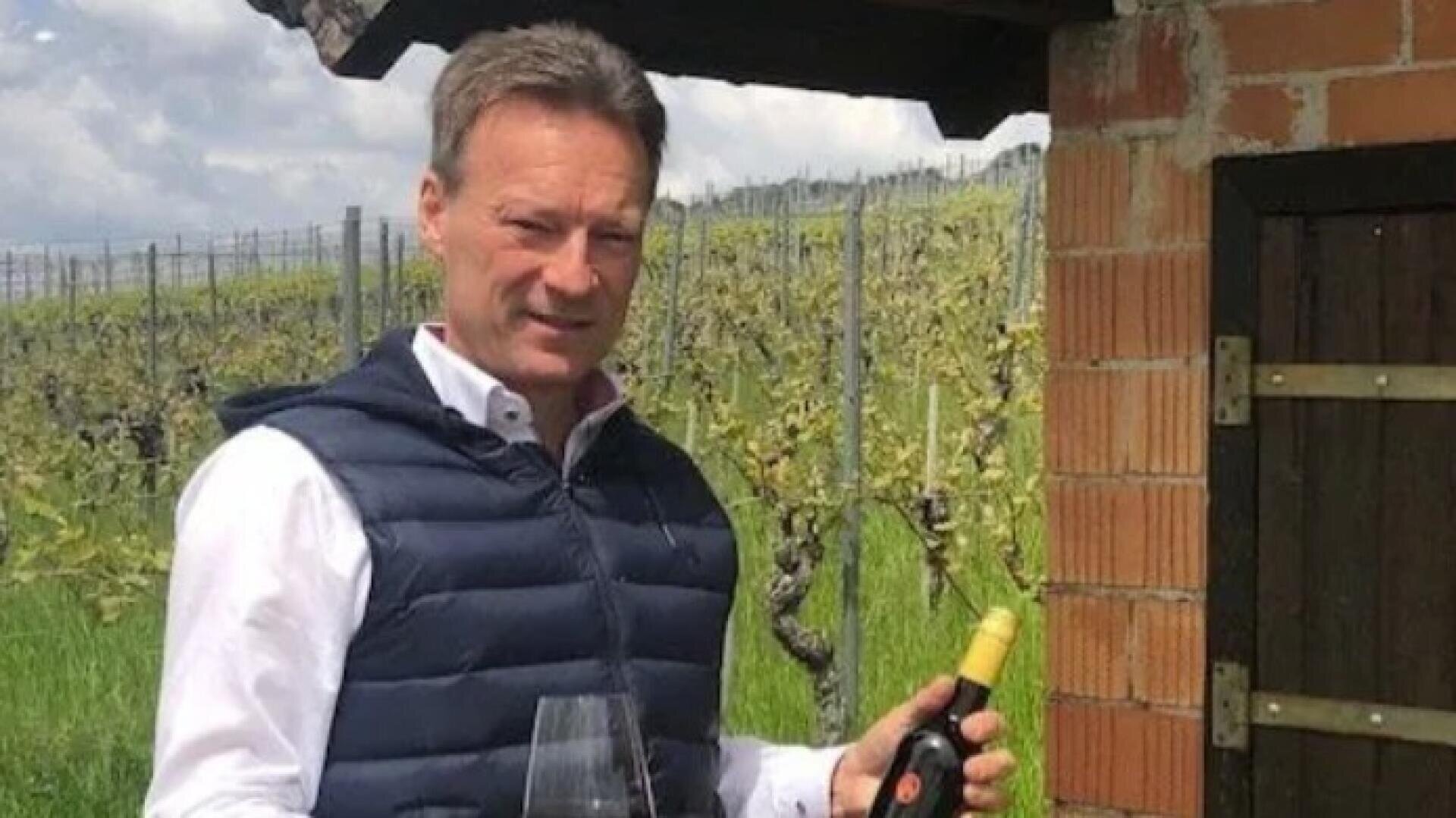 Ein Mann in weißem Hemd und dunkler Weste steht im Freien in einem Weinberg und hält eine Weinflasche und ein Weinglas in der Hand. Im Hintergrund sind grüne Weinreben und ein Backsteingebäude zu sehen.