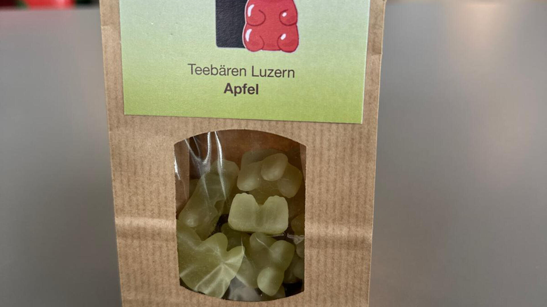 Eine braune Papiertüte mit der Aufschrift Teebären Luzern Apfel enthält grüne Gummibärchen, die durch ein kleines durchsichtiges Fenster am Boden der Verpackung sichtbar sind.