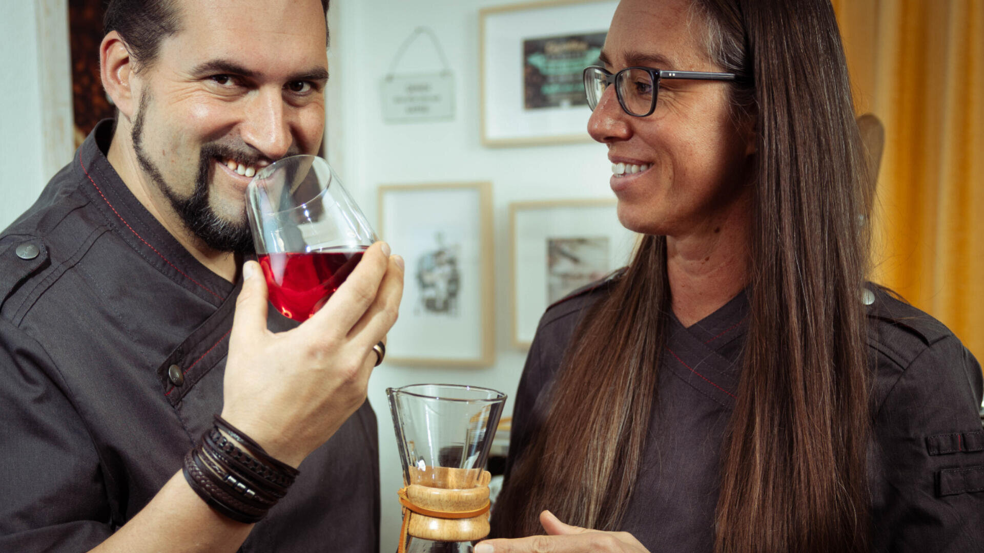 Zwei Personen in dunklen Hemden stehen in einem Innenraum. Eine Person lächelt und schnuppert an einem Glas mit roter Flüssigkeit, während die andere, die eine Brille trägt, eine Kaffeemaschine hält und sie anschaut. An der Wand im Hintergrund hängen gerahmte Bilder.