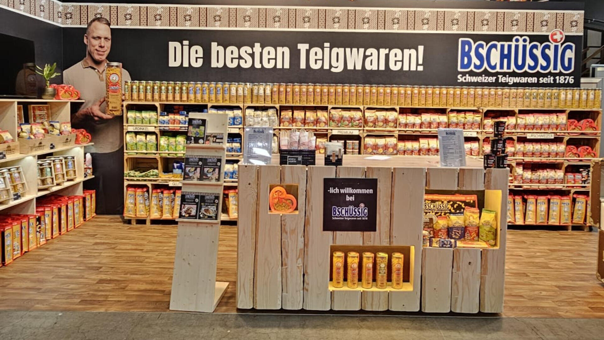 Eine Supermarktauslage für Bschüssig Teigwaren zeigt Regale mit verschiedenen Teigwaren. Im Hintergrund ist ein großes Banner mit einem lächelnden Mann und dem deutschen Text "Die besten Teigwaren!" zu sehen.
