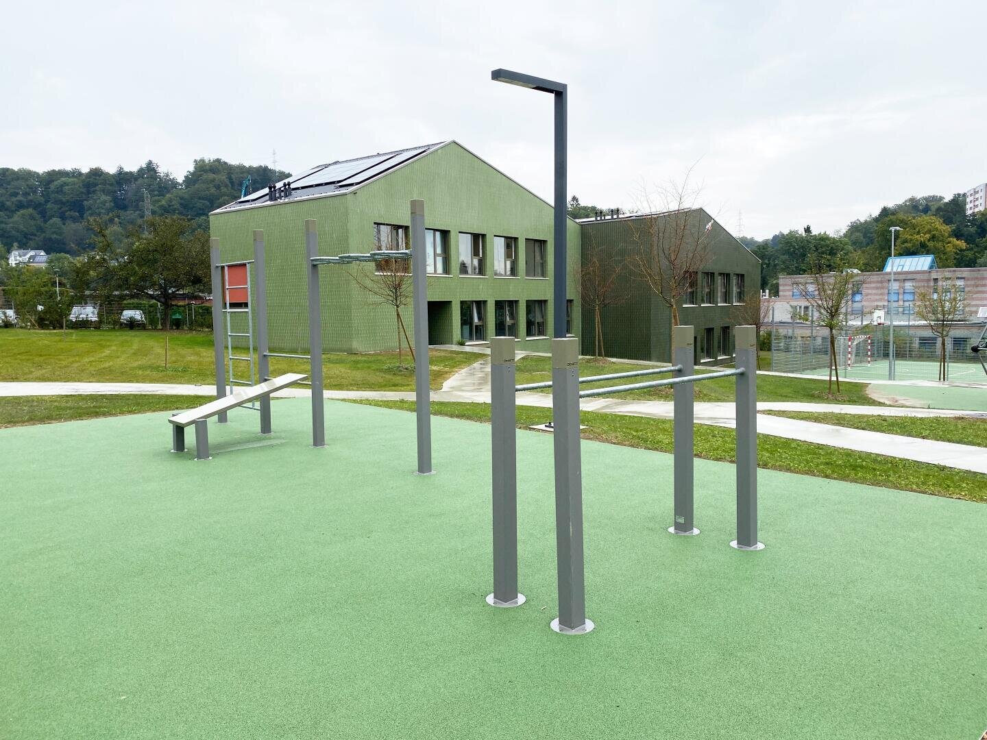 Fitnessbereich im Freien mit Metalltrainingsstäben und einer Bank auf einem grünen Gummibelag, in der Nähe moderner grüner Gebäude und umgeben von Gras und Wegen. Im Hintergrund sind Bäume und Hügel zu sehen.