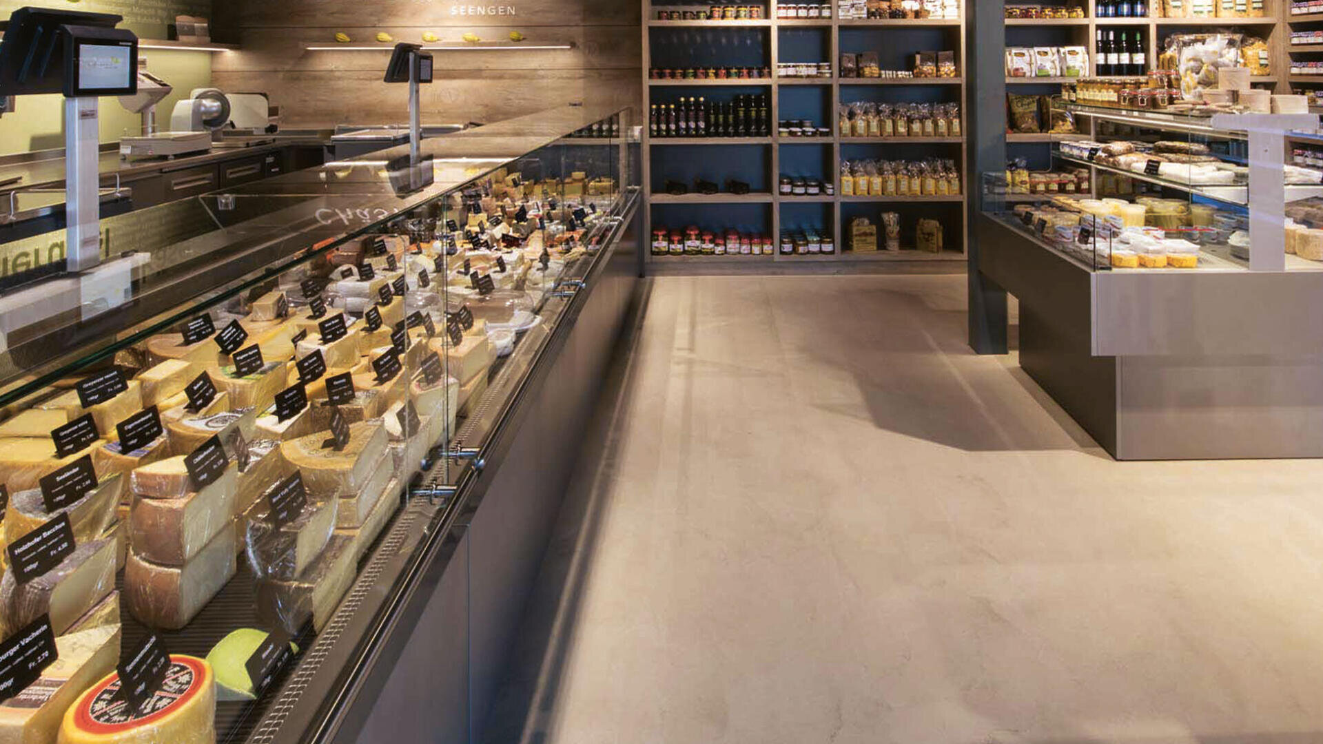 Ein moderner Lebensmittelladen mit einer langen Kühlvitrine mit verschiedenen Käsesorten, Regalen mit verpackten Waren und einer Theke mit verschiedenen Feinkostprodukten. Die Umgebung ist sauber und gut organisiert.