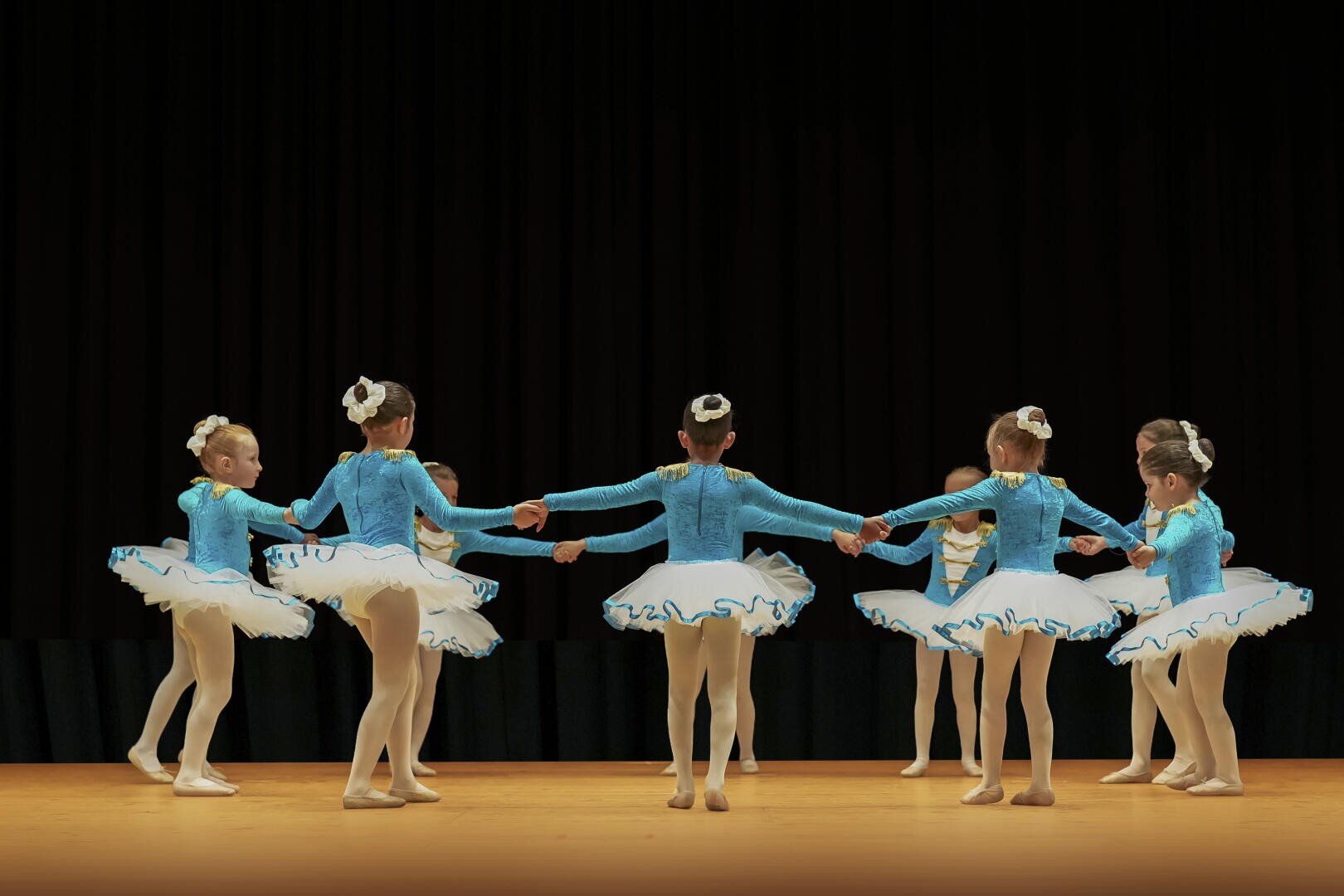 Acht junge Ballerinen in passenden blauen Trikots und weißen Tutus stehen im Kreis und halten sich an den Händen, während sie auf der Bühne eine Tanznummer vor schwarzem Hintergrund aufführen.