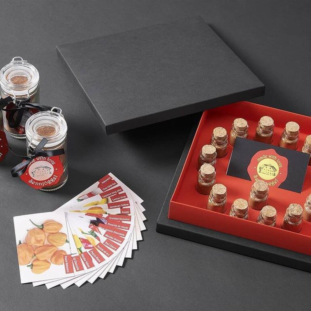 Ein schwarzer Geschenkkarton mit roter Innenseite enthält mehrere kleine verkorkte Fläschchen. Daneben befinden sich Rezeptkarten mit Bildern von Paprika und zwei kleine Gläser mit der Aufschrift "Made with Love". Alle Gegenstände sind auf einer dunklen Fläche angeordnet.