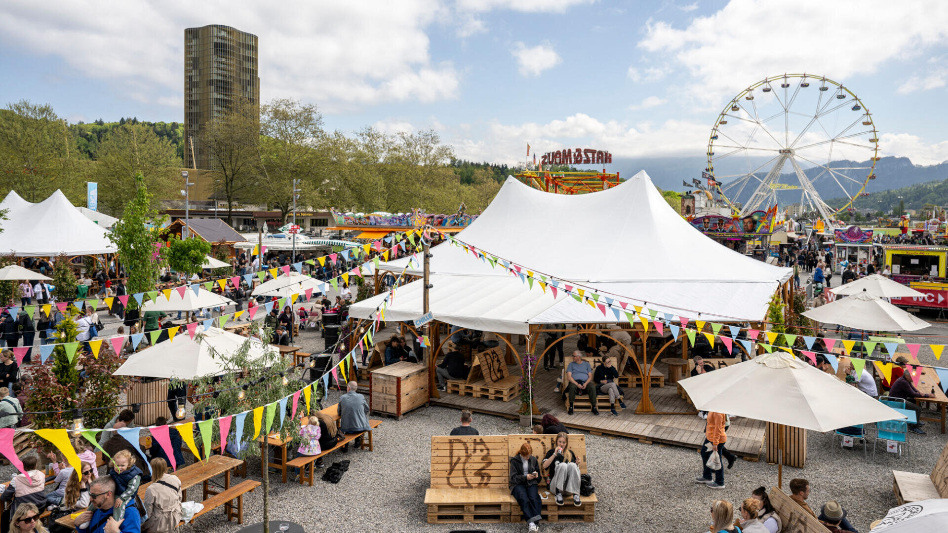 Ein lebhaftes Open-Air-Festival mit bunten Fahnen, weißen Zelten, Essensständen und vielen Menschen, die sitzen und spazieren gehen. Im Hintergrund sind unter einem teilweise bewölkten Himmel ein Riesenrad und ein hohes Gebäude zu sehen.