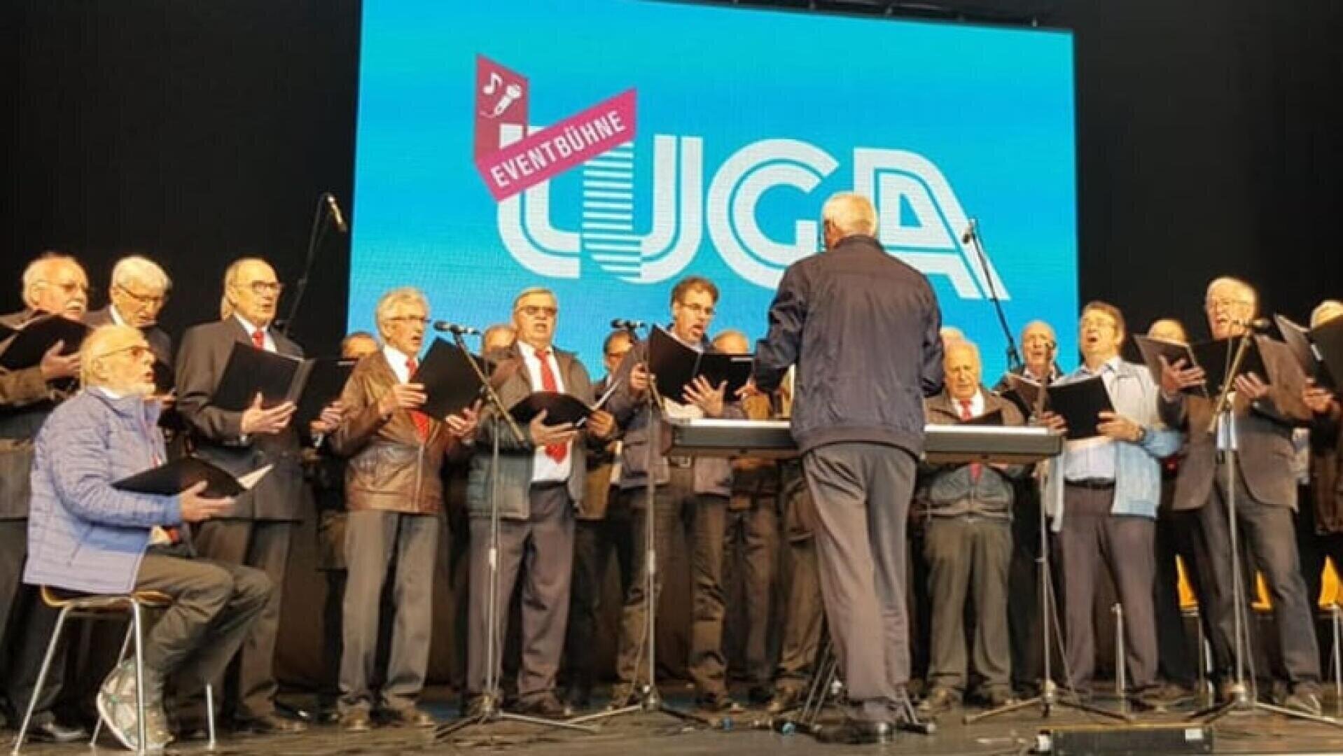 Eine Gruppe älterer Männer singt in einem Chor auf der Bühne, vor ihnen steht ein Dirigent. Hinter ihnen ist ein großer Bildschirm mit dem Wort LUGA und einem rosa Schild mit der Aufschrift EVENTBÜHNE zu sehen.