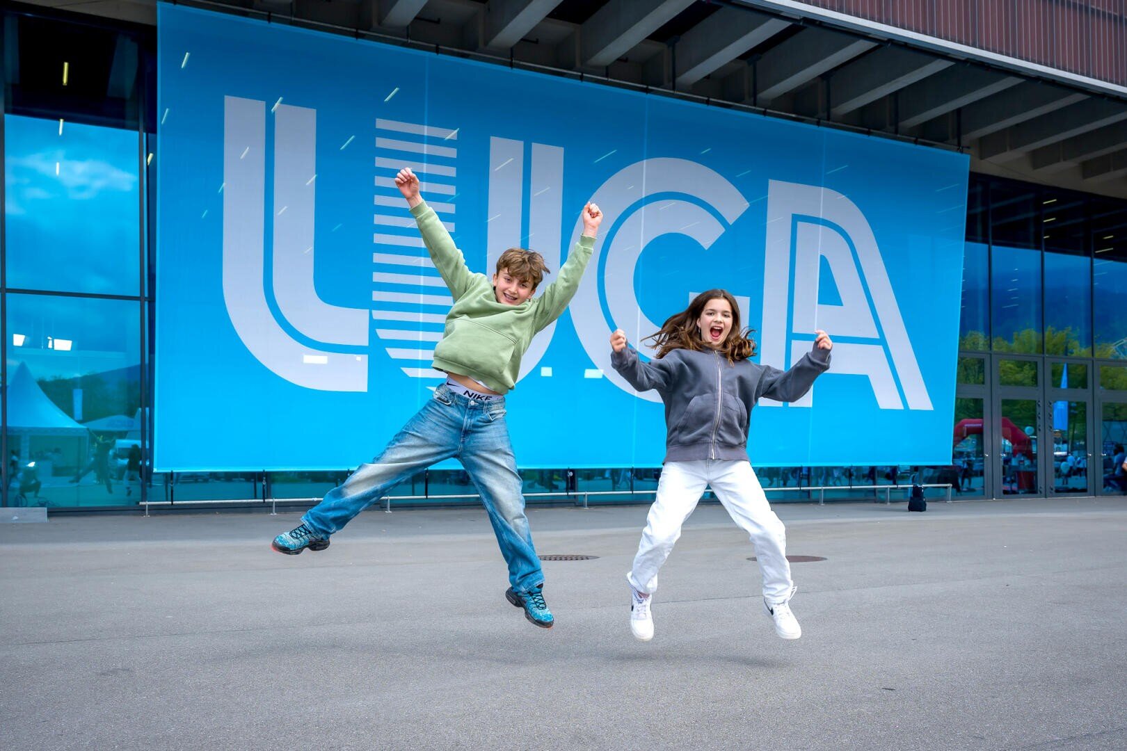 Zwei Kinder in Freizeitkleidung springen vor einem großen blauen Banner mit der Aufschrift LUCA vor einem modernen Gebäude mit Glastüren und -fenstern fröhlich herum.