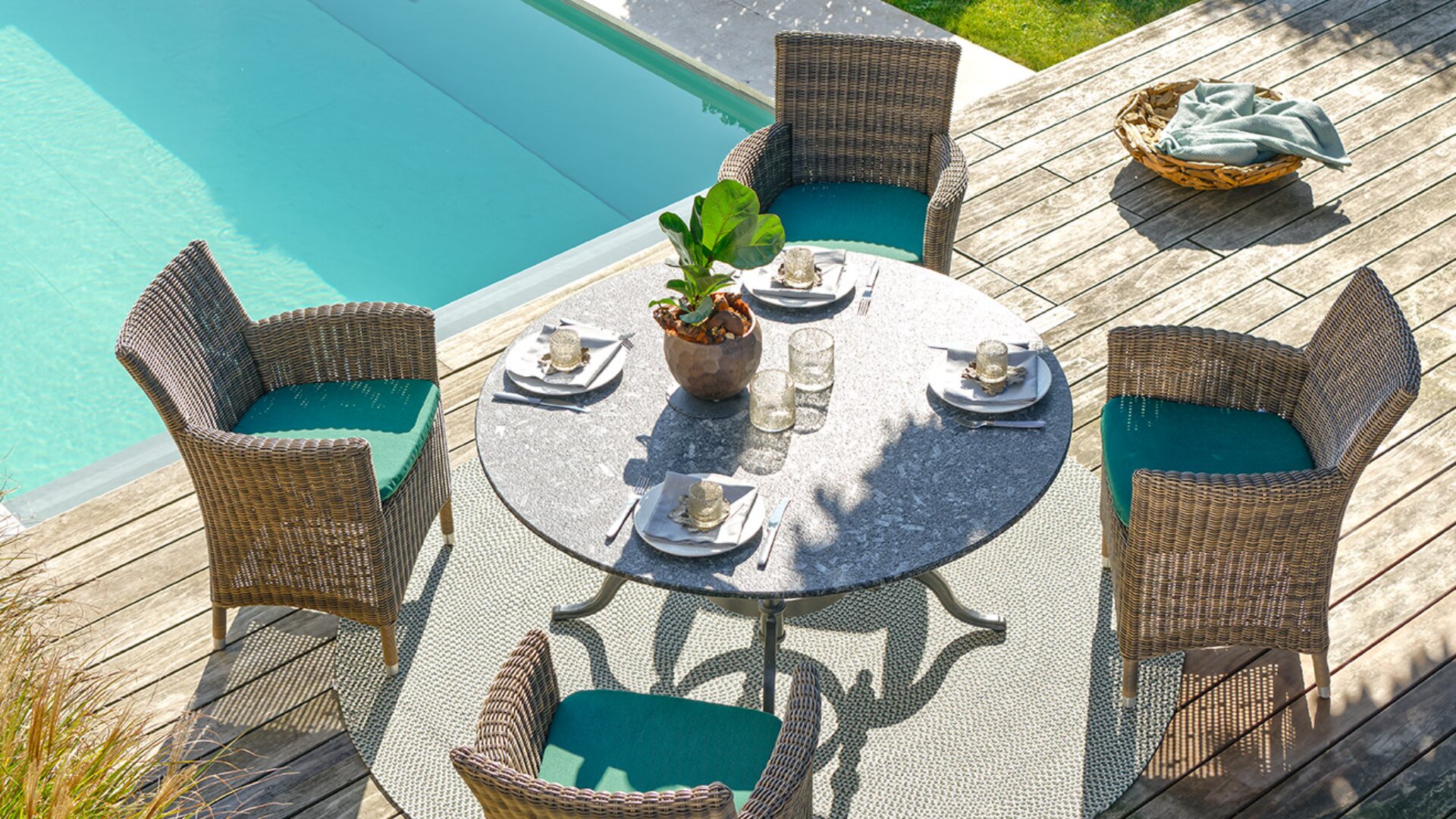 Ein runder Tisch mit vier Korbstühlen steht auf einer Holzterrasse an einem Swimmingpool. Der Tisch ist mit Tassen, Tellern und Servietten gedeckt, und eine Topfpflanze dient als Mittelstück. Das Sonnenlicht wirft Schatten auf die Szene.