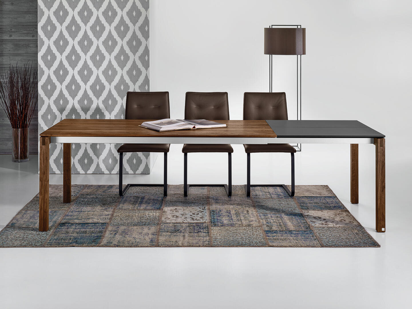 Ein modernes Esszimmer mit einem rechteckigen Tisch aus Holz und Schwarz, drei braunen Stühlen, einer Stehlampe, einer Vase mit hohen Zweigen und einer geometrisch gemusterten Wand mit einem Flickenteppich unter dem Tisch.