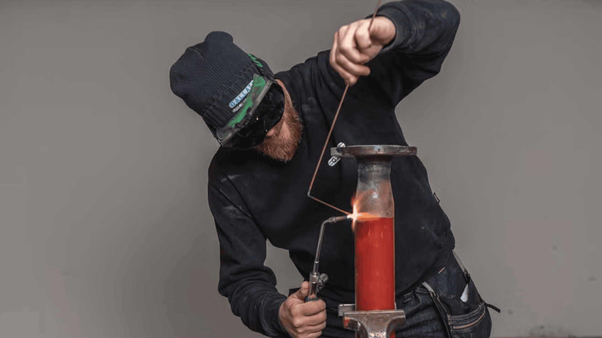 Eine Person, die eine Schutzbrille, eine Mütze und dunkle Kleidung trägt, schweißt oder schneidet mit einem Schweißbrenner ein Metallrohr, wobei an der Kontaktstelle leuchtend orangefarbene Flammen zu sehen sind.