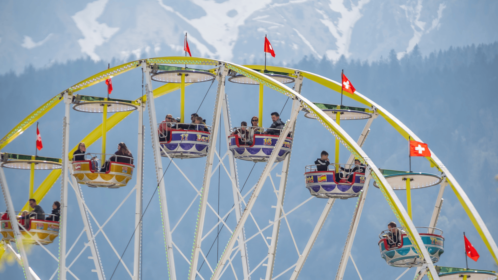 Ein farbenfrohes Riesenrad mit Menschen in den Gondeln, die mit Schweizer Flaggen geschmückt sind, steht vor einer Kulisse aus verschneiten Bergen und Kiefern.