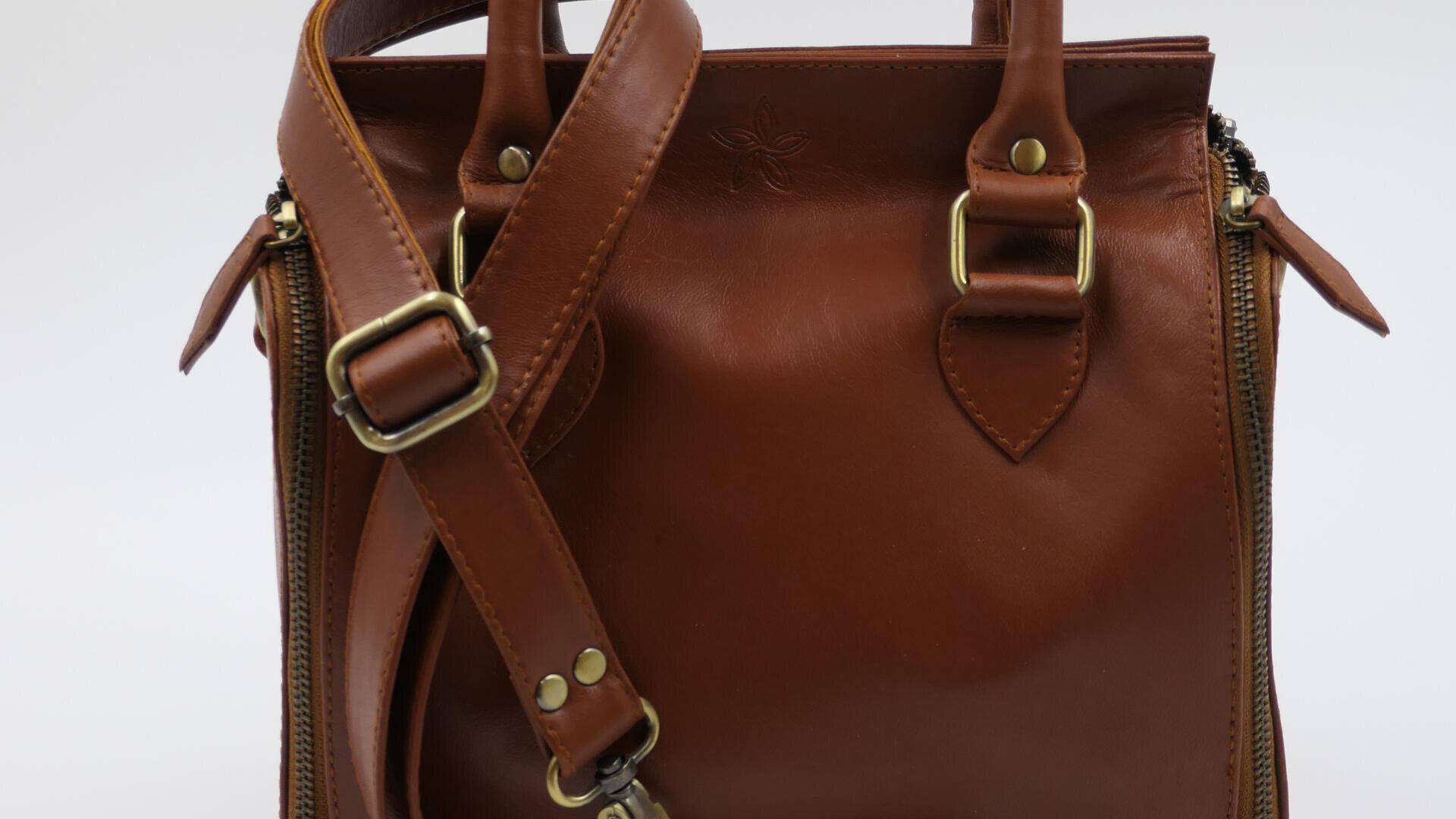 Eine braune Lederhandtasche mit goldenen Schnallen, einem Schulterriemen und zwei Griffen. Die Tasche hat eine glatte Oberfläche und sichtbare Nähte, mit Reißverschlüssen auf beiden Seiten.