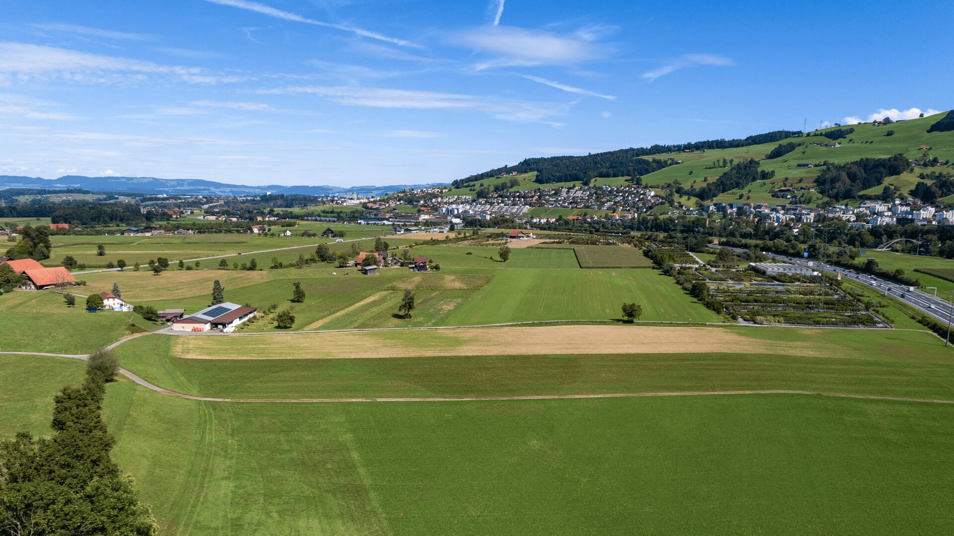 Luftaufnahme einer ländlichen Landschaft mit grünen Feldern, verstreuten Häusern und einer kleinen Stadt im Hintergrund unter einem blauen Himmel mit Wolkenfetzen. Sanfte Hügel und Baumgruppen umgeben das Gebiet.