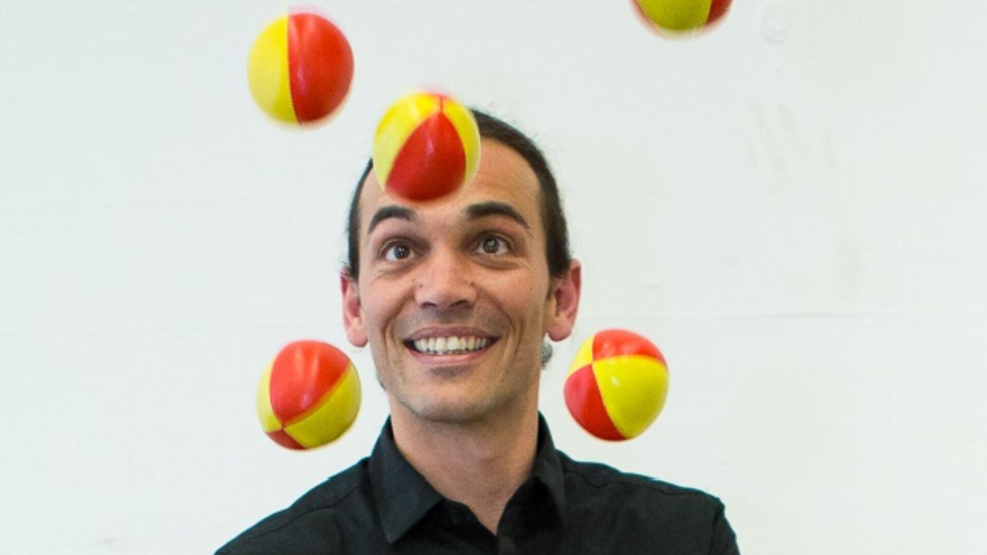 Eine lächelnde Person in einem schwarzen Hemd jongliert sechs rote und gelbe Bälle vor einem schlichten weißen Hintergrund.