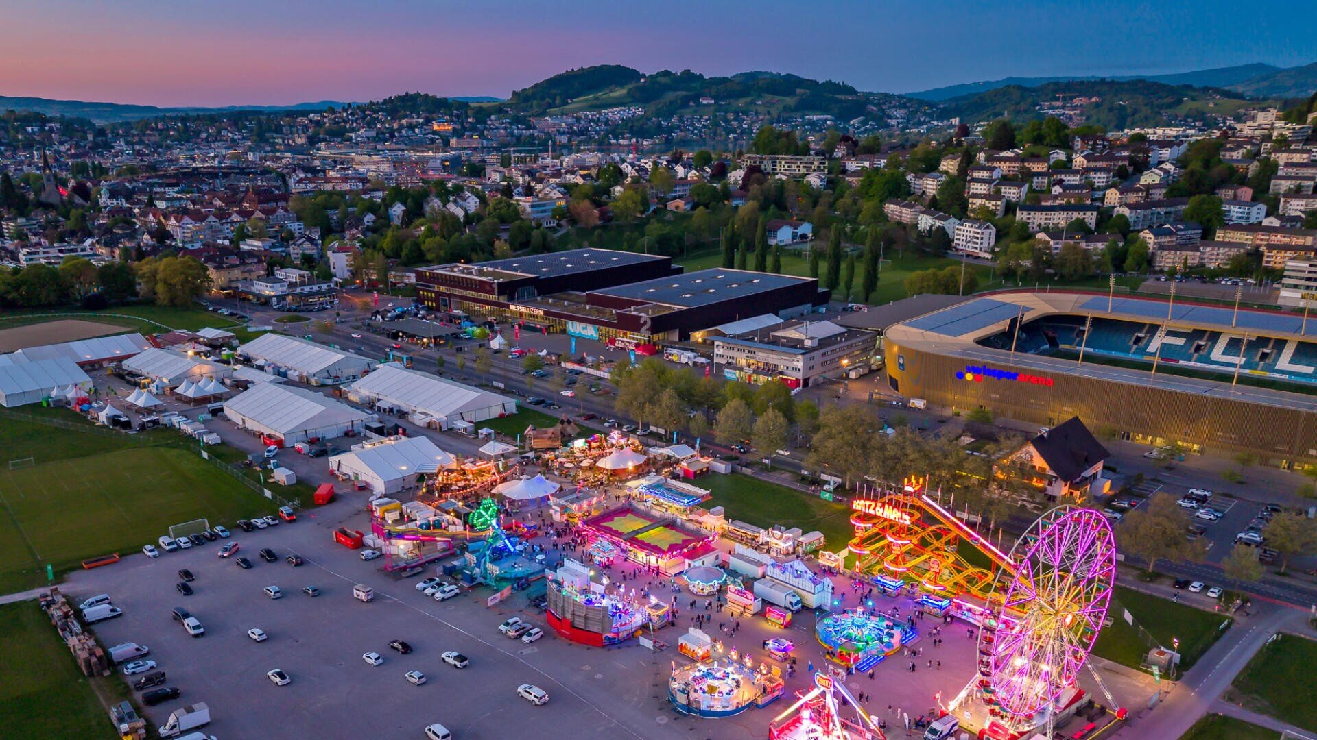 Luftaufnahme eines lebhaften Jahrmarkts mit bunten Fahrgeschäften und Attraktionen, umgeben von geparkten Autos und Zelten. Im Hintergrund ist bei Sonnenuntergang eine Stadt mit Hügeln und Häusern zu sehen.