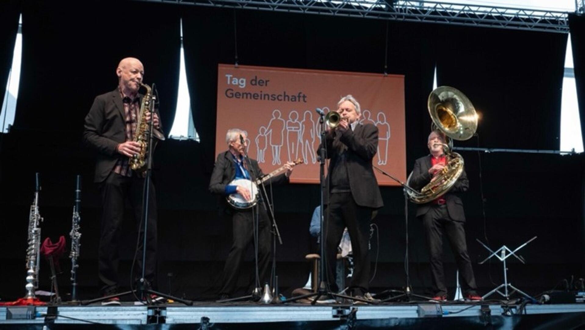 Vier Musiker spielen auf der Bühne mit Saxophon, Banjo, Posaune und Sousaphon vor einem Schild mit der Aufschrift "Tag der Gemeinschaft". Schwarzer Hintergrund und Notenständer sind sichtbar.